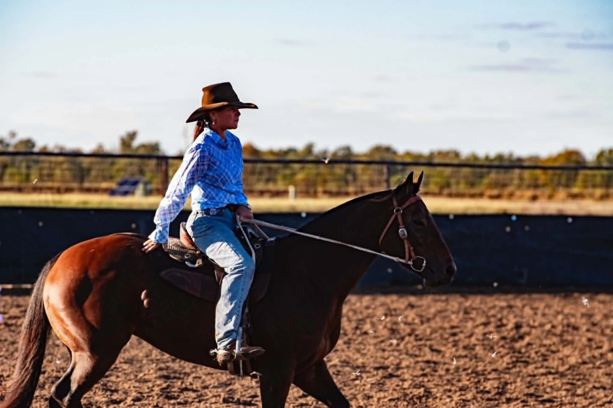 A person wearing a cowboy hat and a blue checked shirt riding a brown horse on a dirt track during daytime.