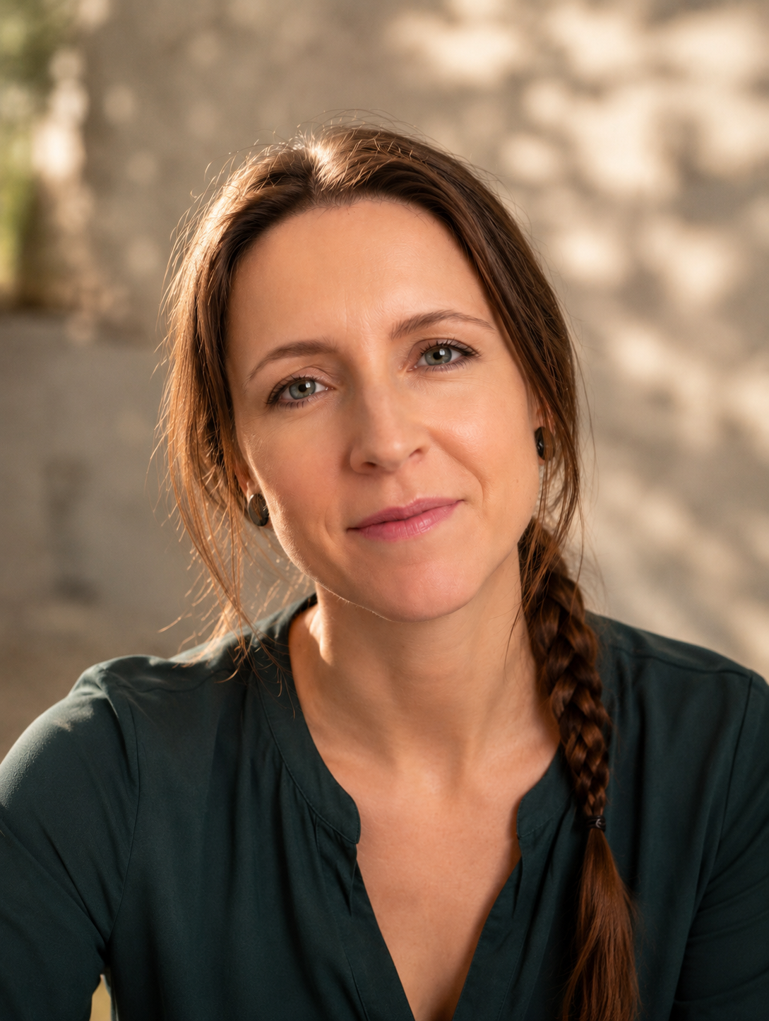 Nahaufnahme einer Frau mit braunem Haar, das zu einem Zopf geflochten ist, trägt schwarze Kleidung und kleine schwarze Ohrringe, vor einem unscharfen grauen Hintergrund.