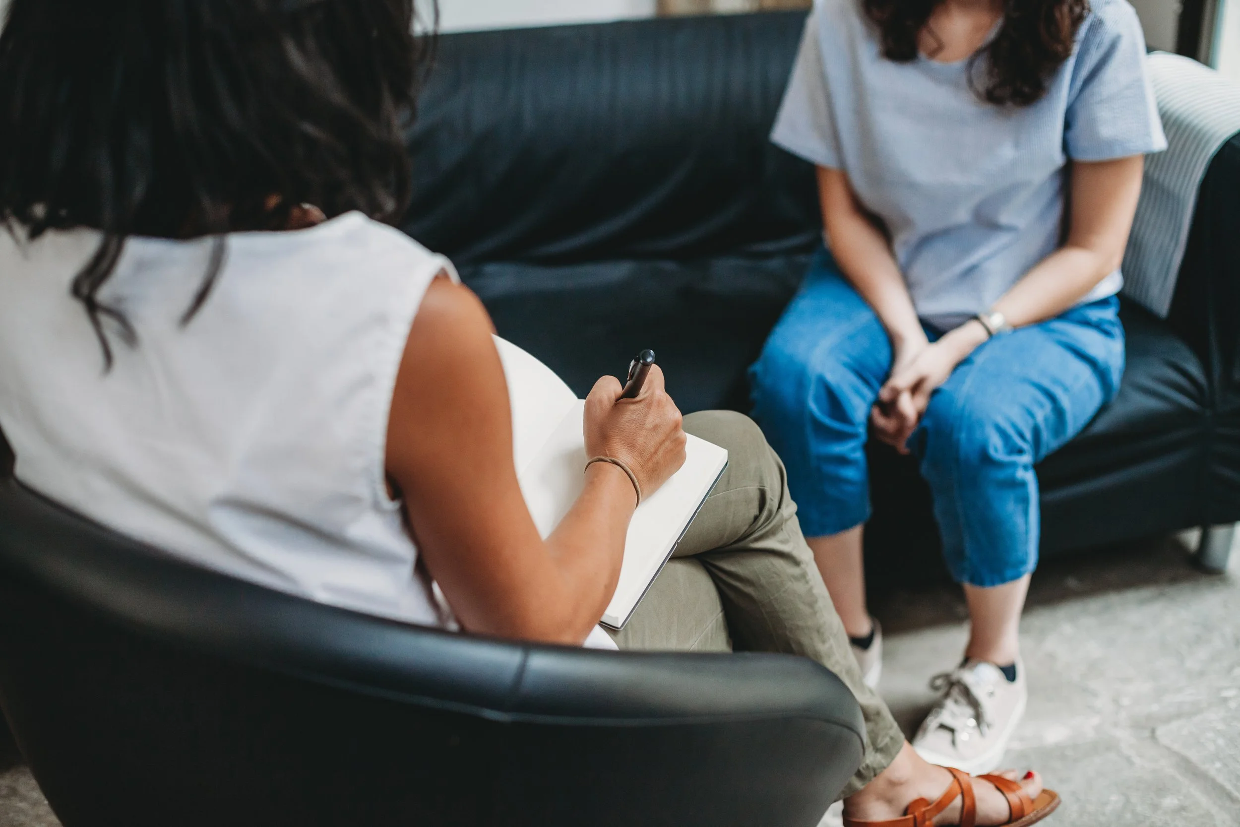 Zwei Frauen sitzen auf einer Couch, eine schreibt in ein Notizbuch, die andere sitzt gegenüber, trägt Jeans und weiße Schuhe, im Hintergrund eine graue Wand.