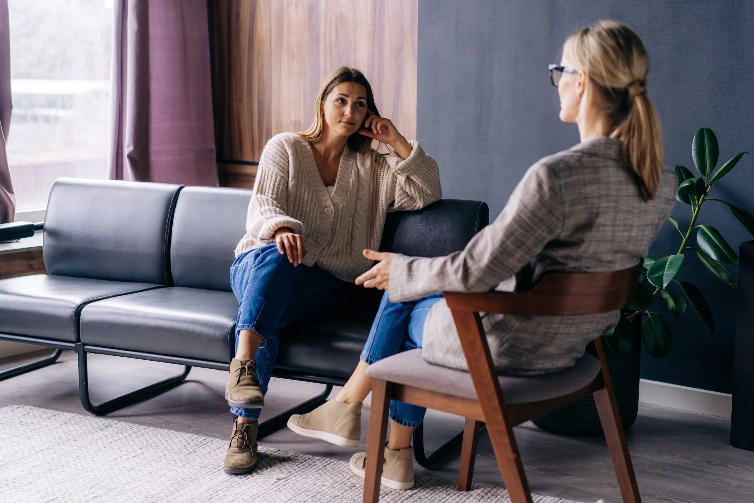 Zwei Frauen unterhalten sich in einem modernen, hellen Raum mit viel natürlichem Licht, einer mit braunen Haaren auf einem schwarzen Sofa, die andere mit blonden Haaren in einem Stuhl.