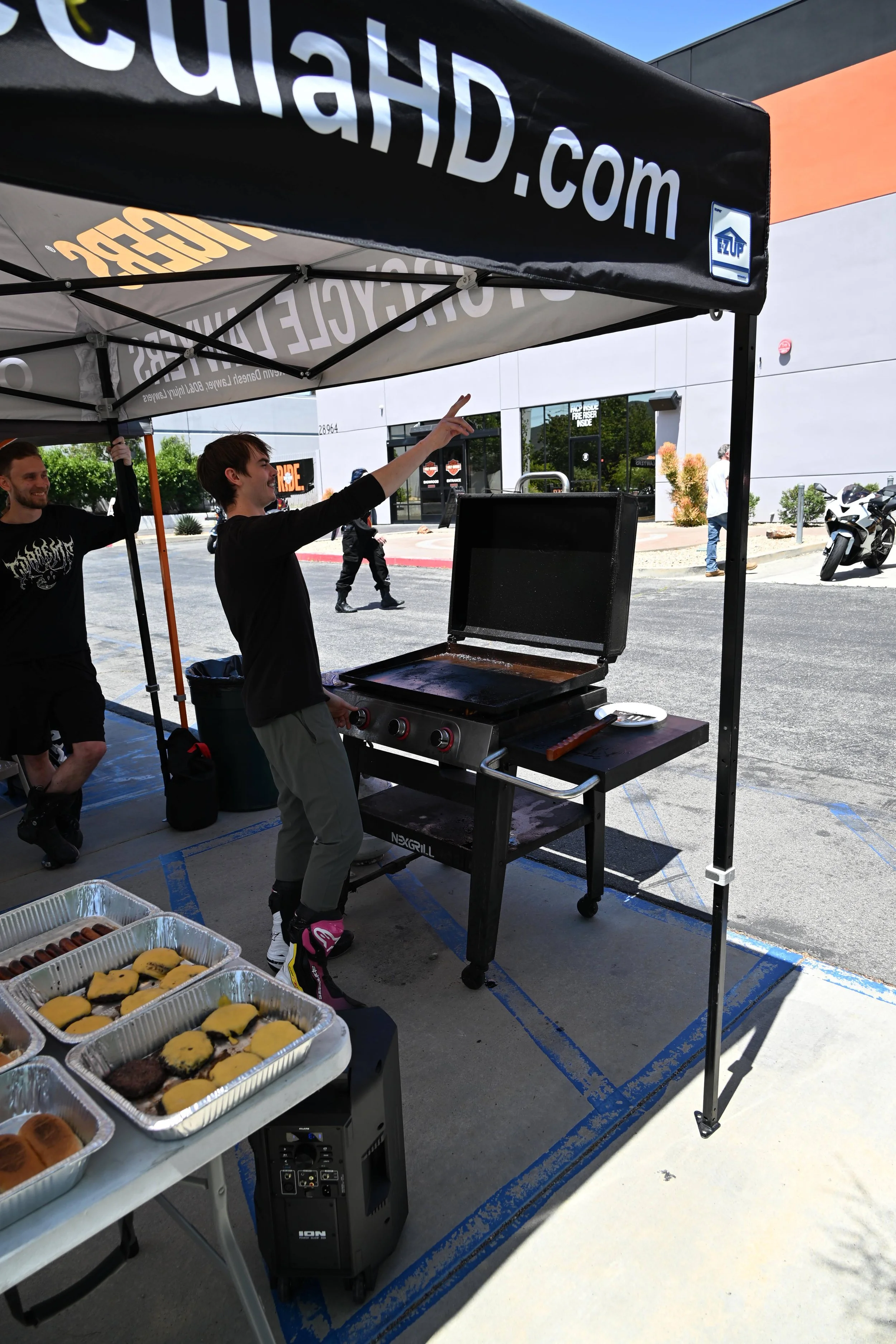 People preparing food at a barbecue stand outdoors with a grill, snacks on display, and a large tent with advertising.