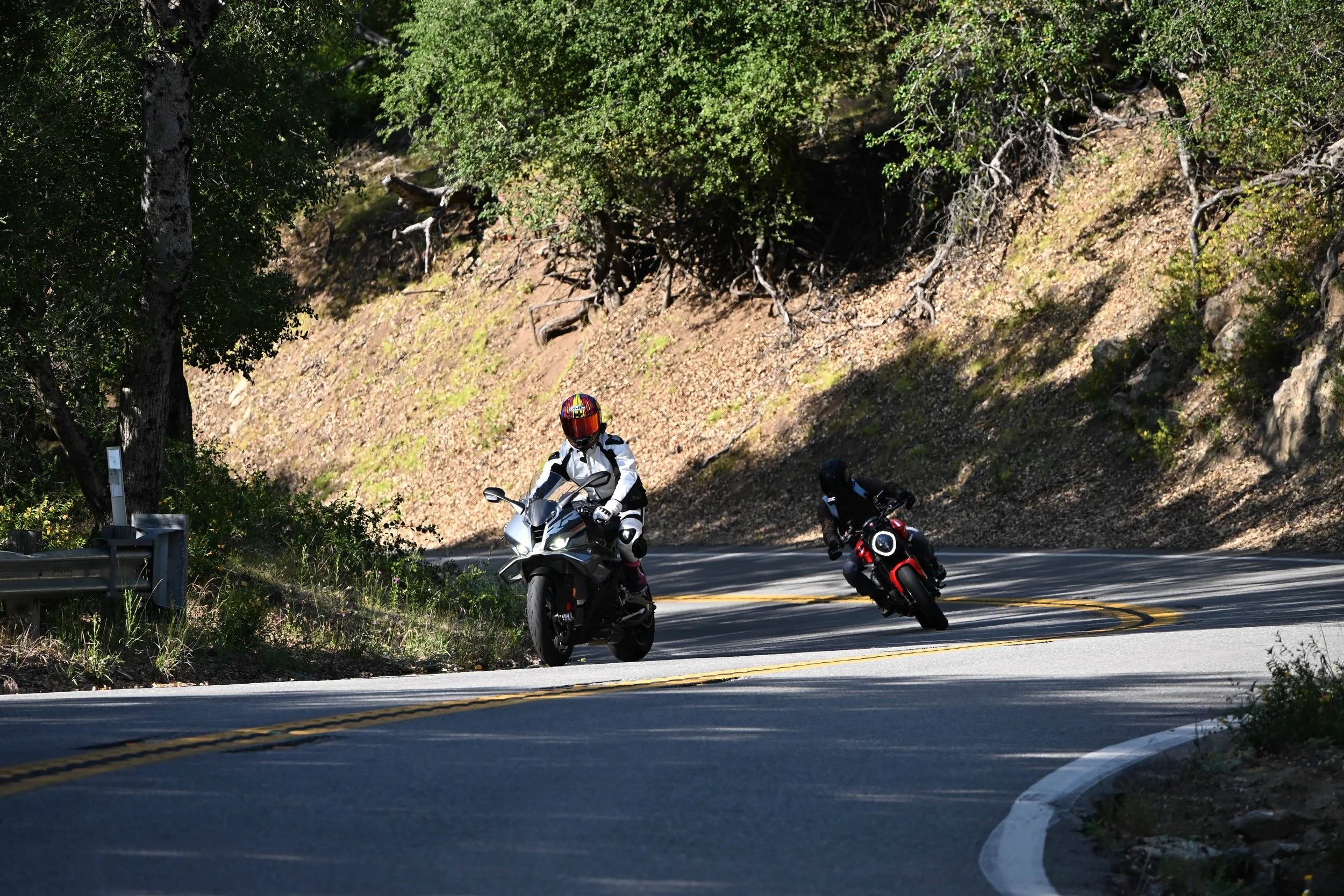 Two motorcyclists riding on a winding mountain road surrounded by trees and rocky terrain.