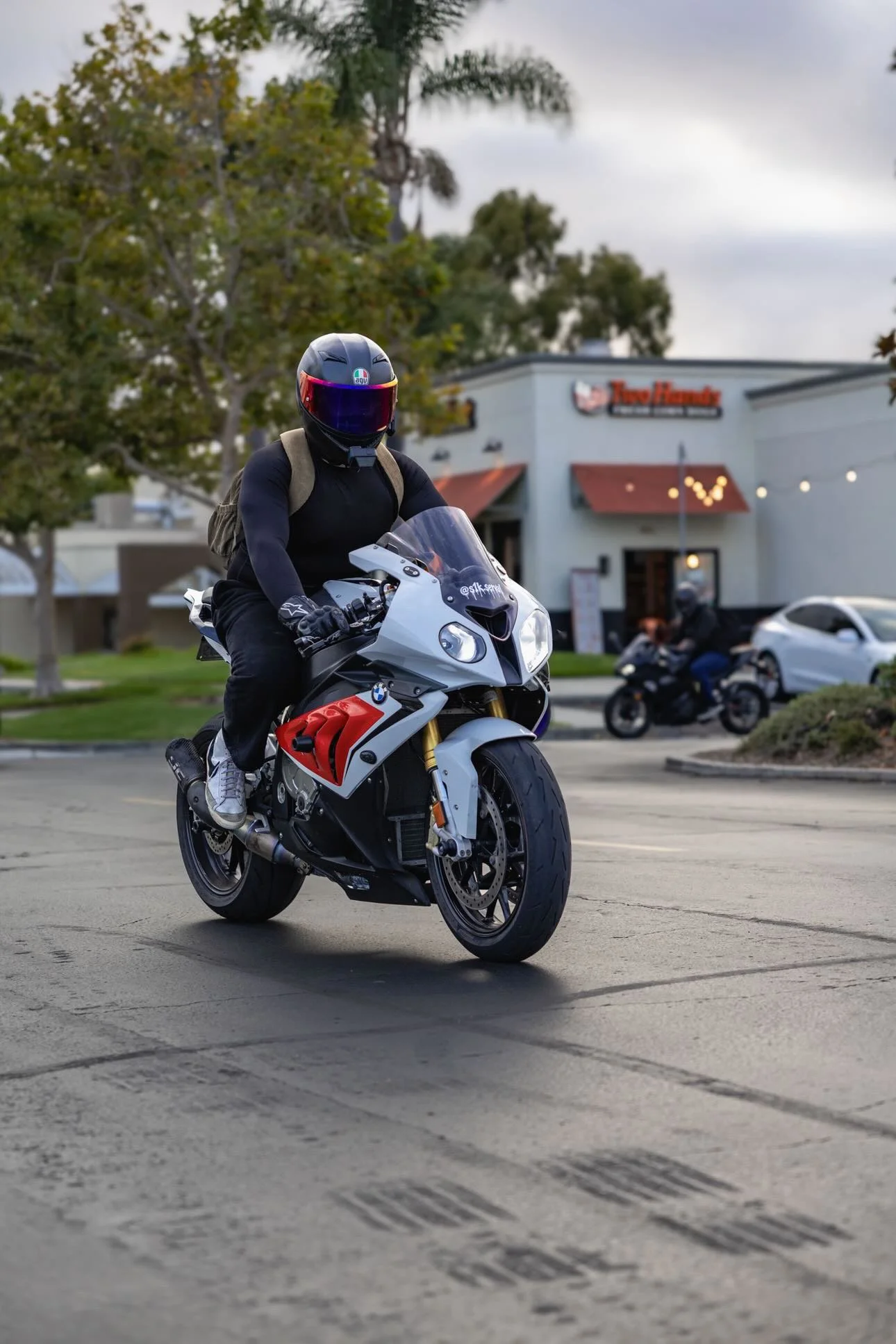 A person wearing a black helmet, black jacket, and gloves rides a white BMW sport motorcycle on a paved lot with tire marks. There are trees and a building with a sign reading "Tualatin" in the background, and a cloudy sky overhead.