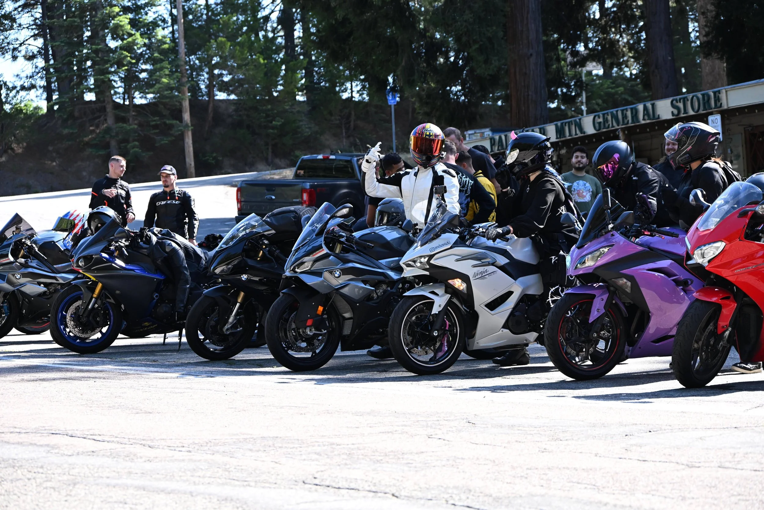 A group of motorcyclists gathered together, with several sport bikes parked in front of them near a general store in a forested area.