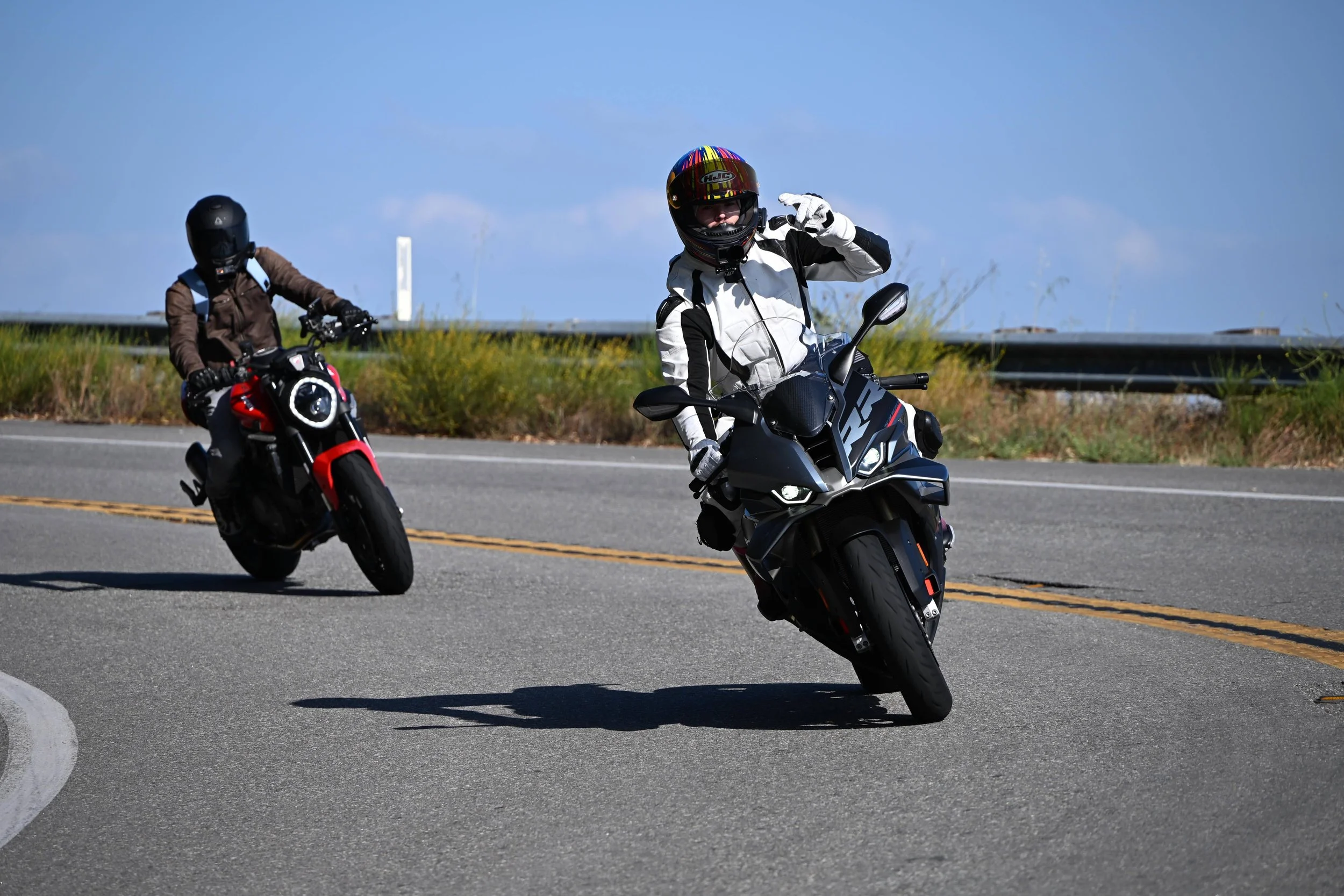 Two motorcyclists riding on a highway, one in a white and black suit with a colorful helmet, and the other in darker clothing with a black helmet, with a clear blue sky and roadside vegetation.