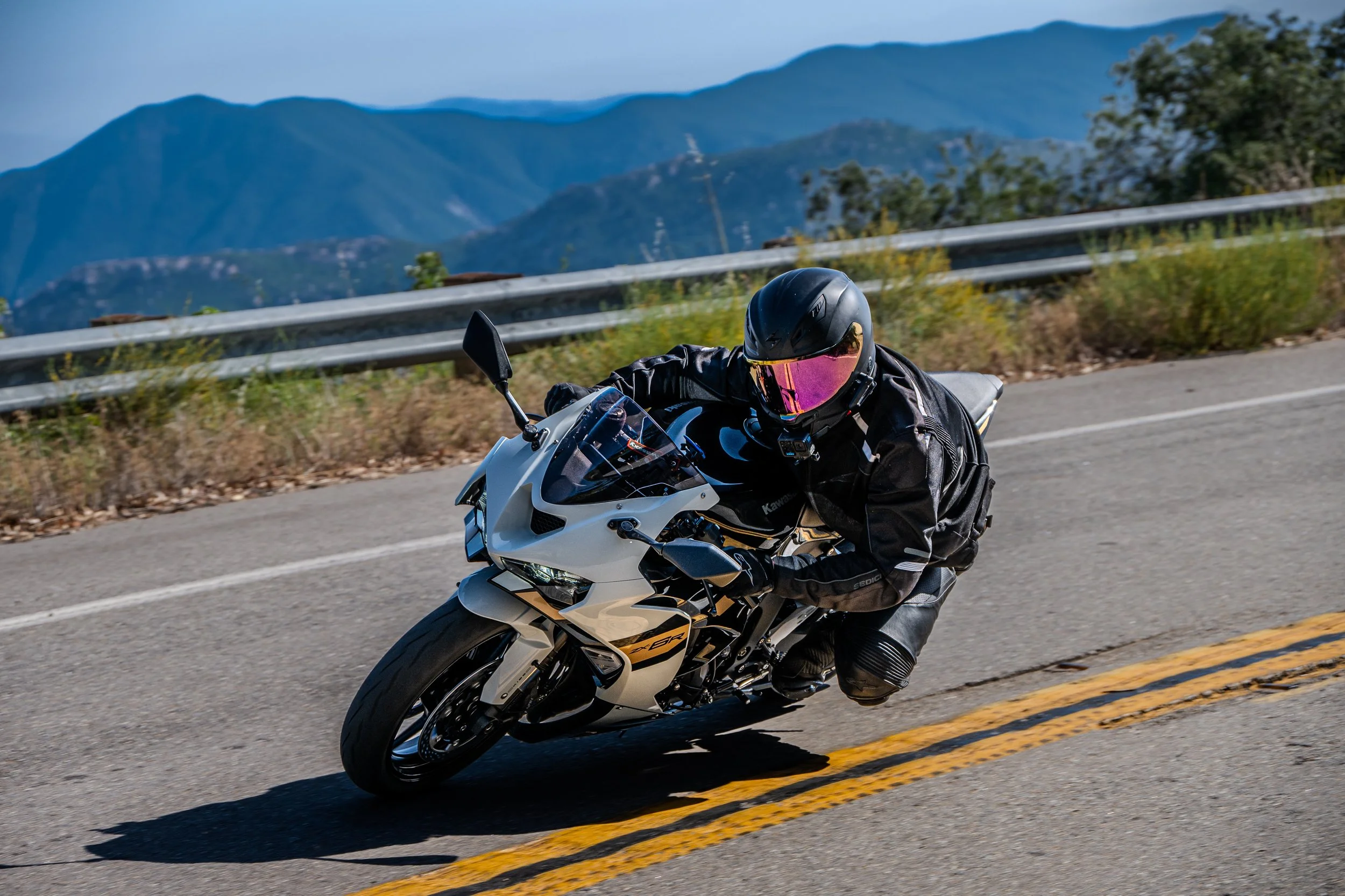 A motorcyclist in black gear and a black helmet with a pink visor riding a white motorcycle on a mountain road with mountains and blue sky in the background.