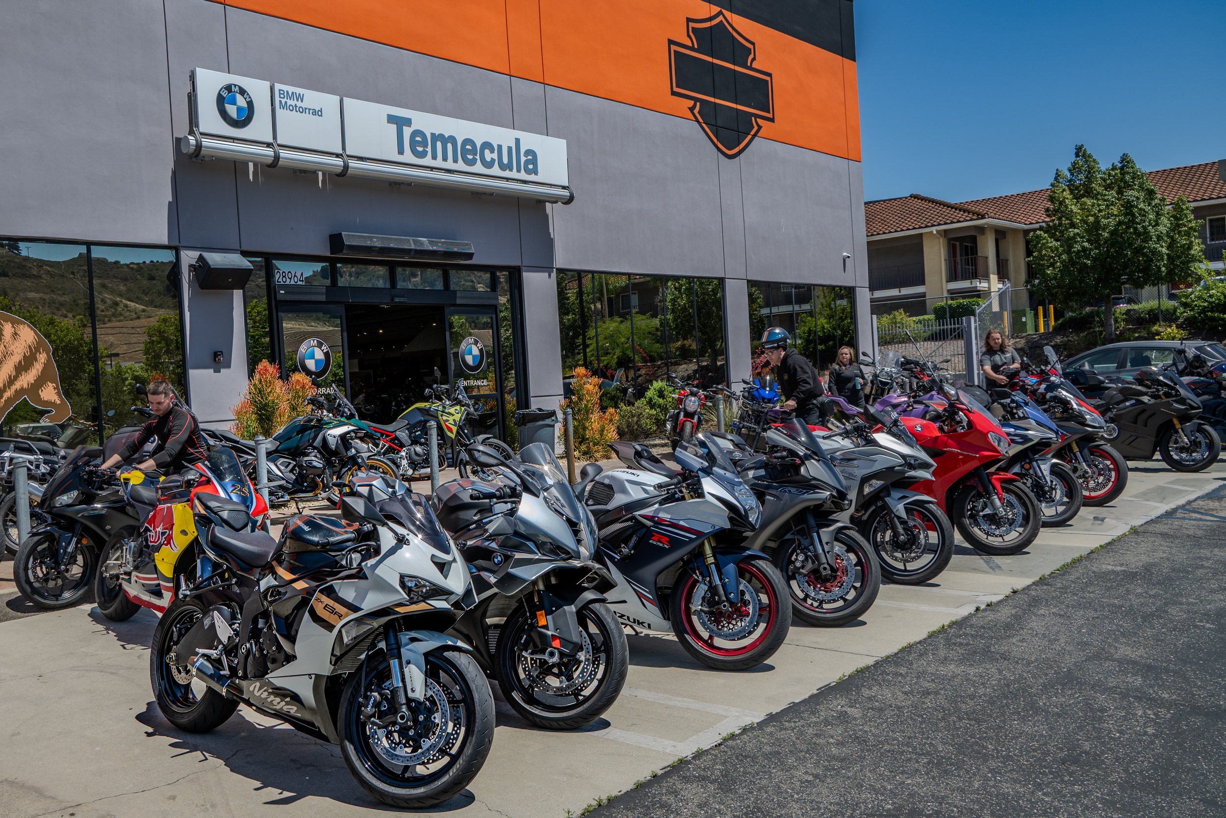 A parking lot filled with motorcycles in front of a motorcycle dealership, with a large sign that says 'Temecula' and the BMW Motorrad logo. There are several people near the motorcycles and a building with residential apartments in the background.
