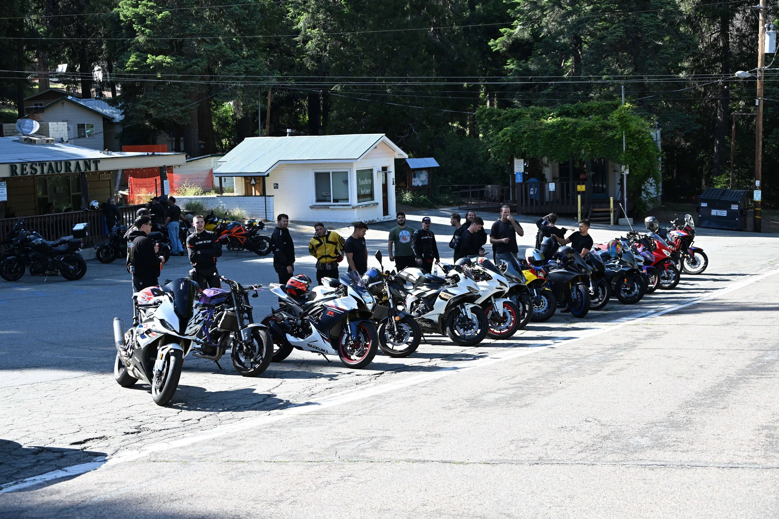 Motorcycles parked in a lot with a group of people standing nearby, some talking and others looking at the bikes, next to small buildings and surrounded by trees.