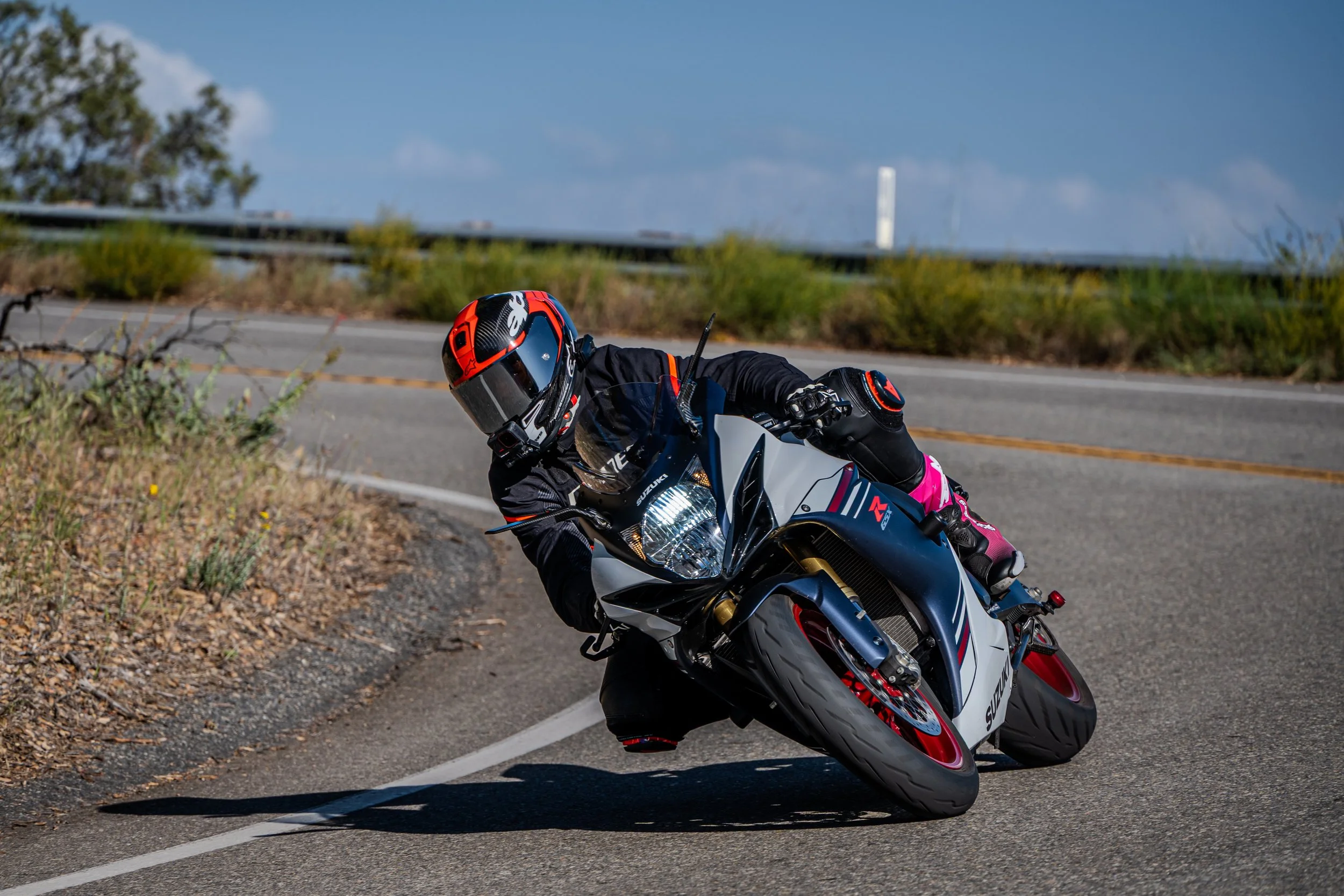 Motorcycle rider in black and pink gear leaning into a turn on a paved road with a guardrail and natural landscape in the background.