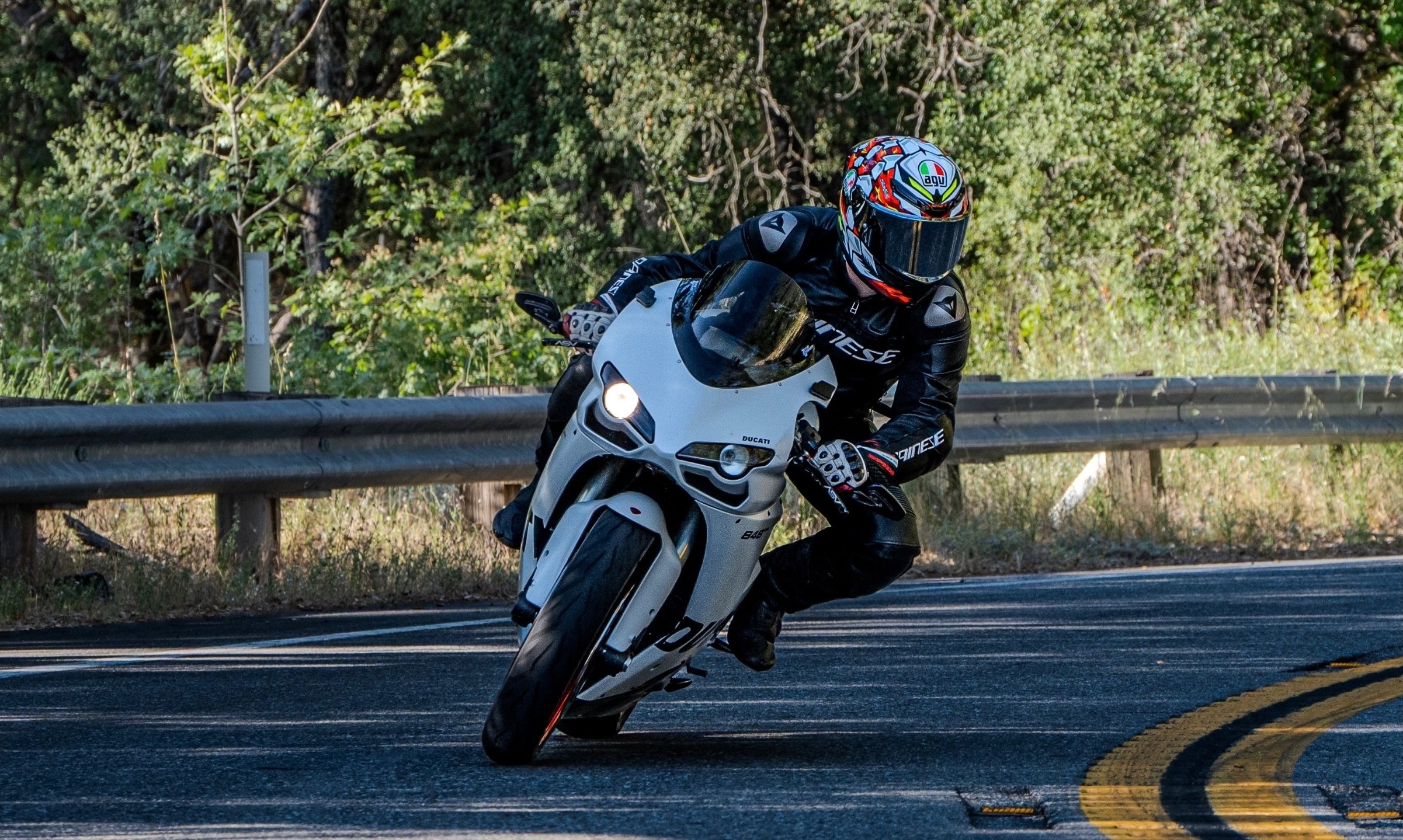 Motorcycle rider wearing black gear and a colorful helmet leaning into a turn on a curved road surrounded by green trees.