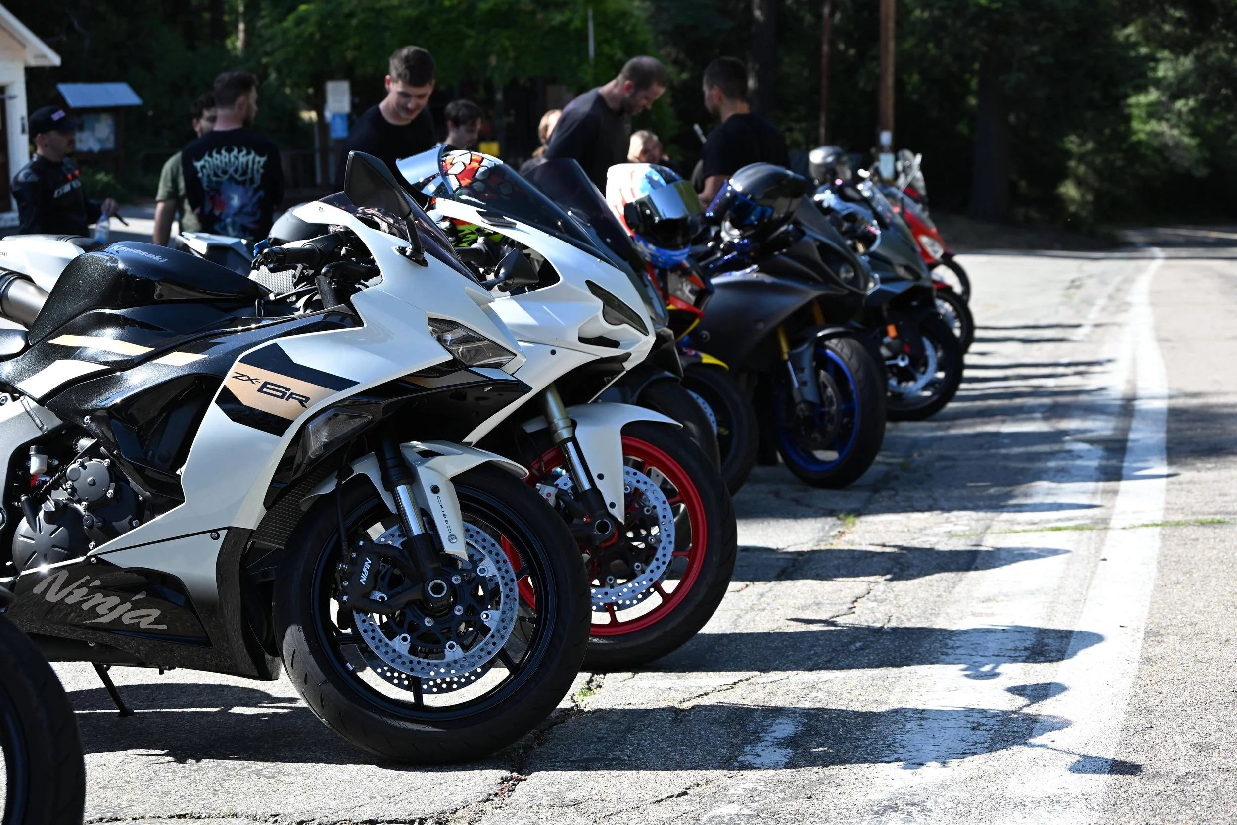 Line of sport motorcycles parked on a concrete surface with people standing behind them, outdoors with trees in the background.