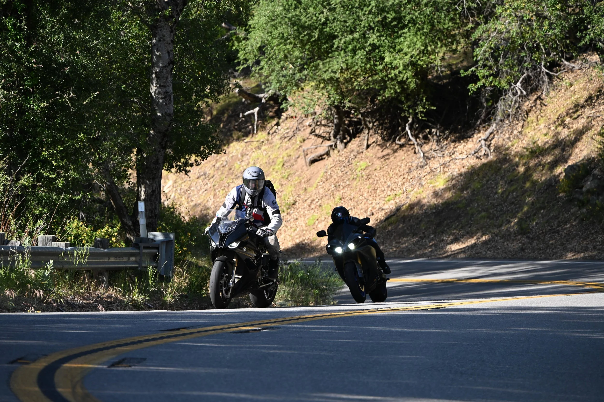 Two motorcyclists riding along a curvy mountain road surrounded by tall green trees and rocky terrain.