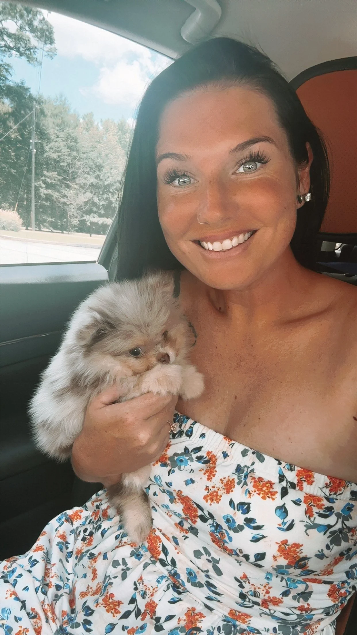 A woman with dark hair and light eyes sitting in a vehicle, smiling, and holding a fluffy puppy with light fur and darker markings.