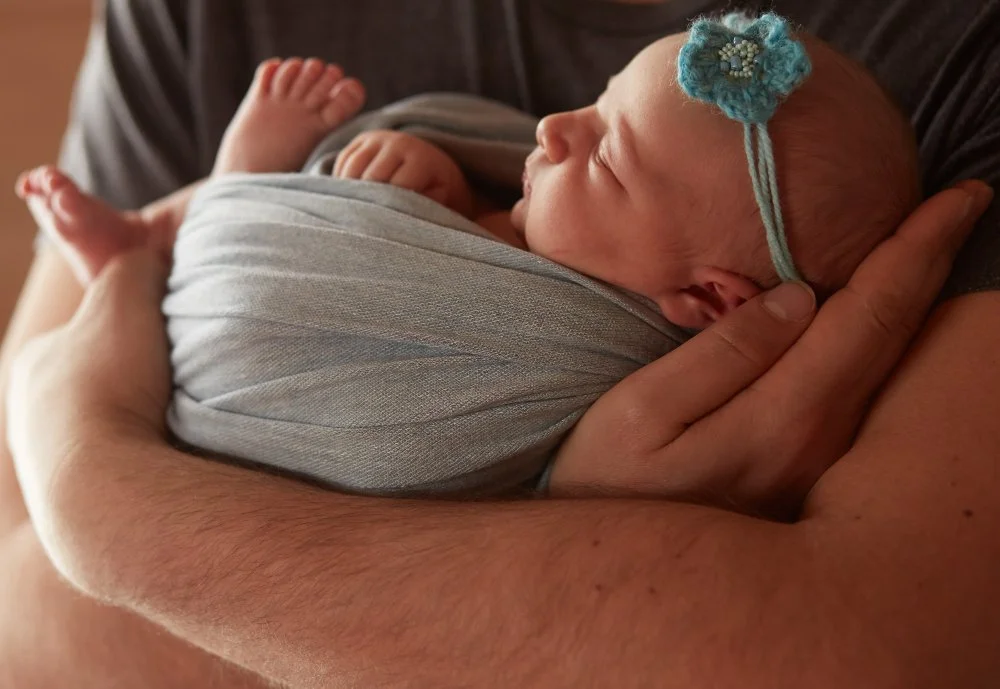 Newborn baby wrapped in a gray blanket, lying in an adult's arms, with a small blue headband with a flower.
