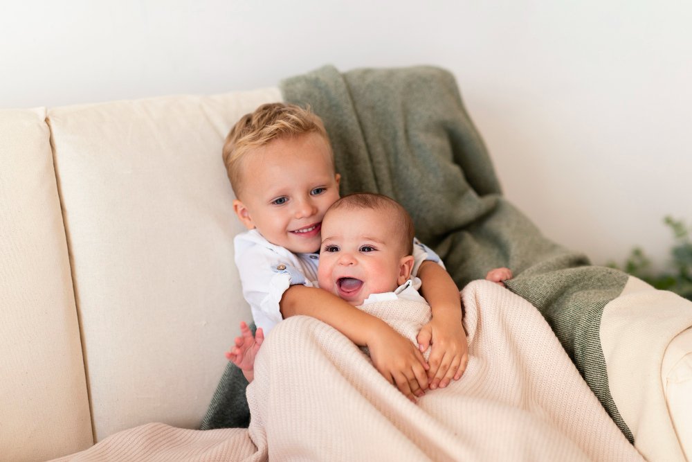 Two young children, a boy and a baby, sitting on a beige couch with a green blanket, smiling and hugging each other.