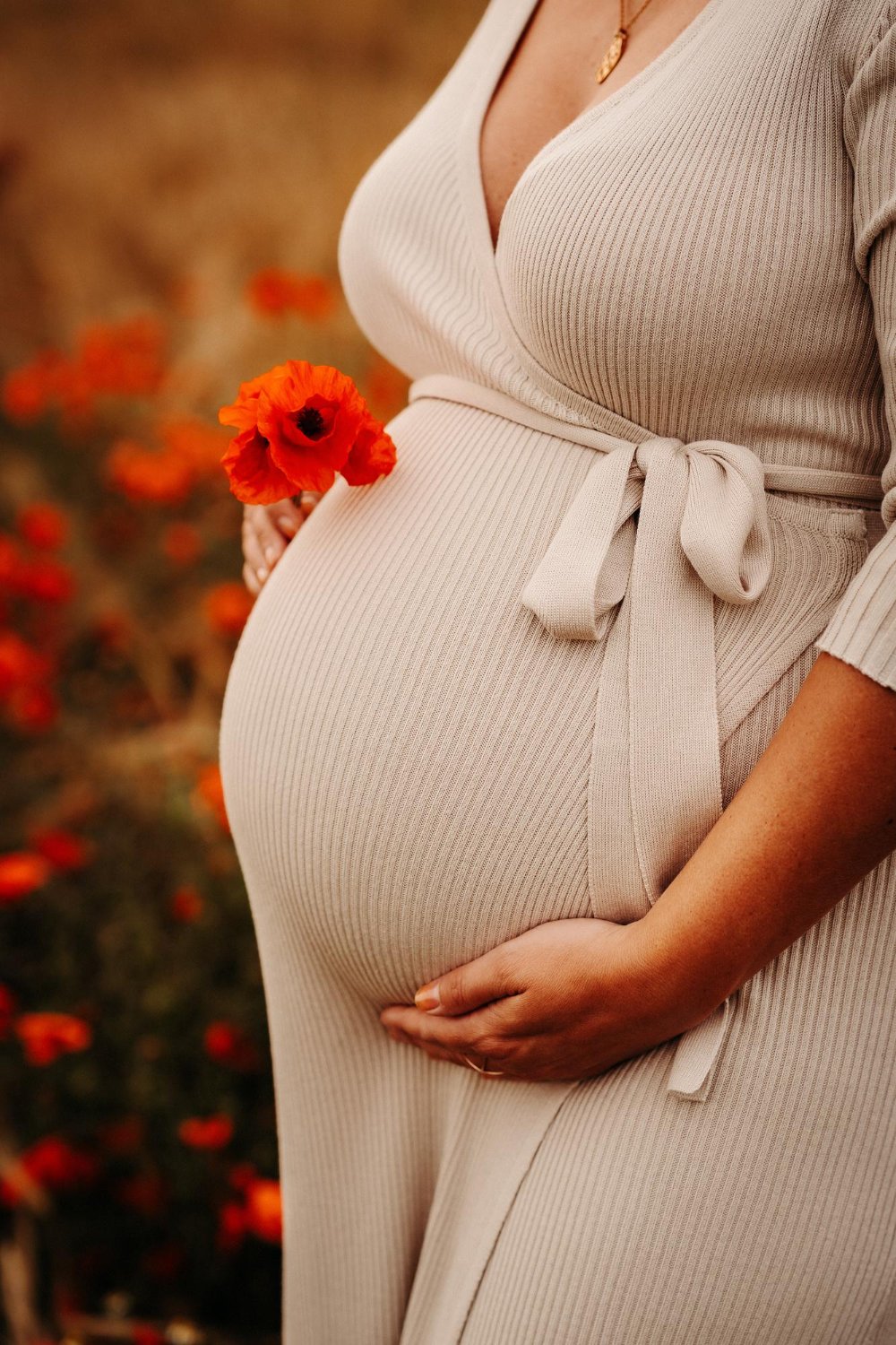 Close-up of a pregnant woman in a beige ribbed dress holding an orange poppy flower near her belly, with a blurred background of similar flowers.