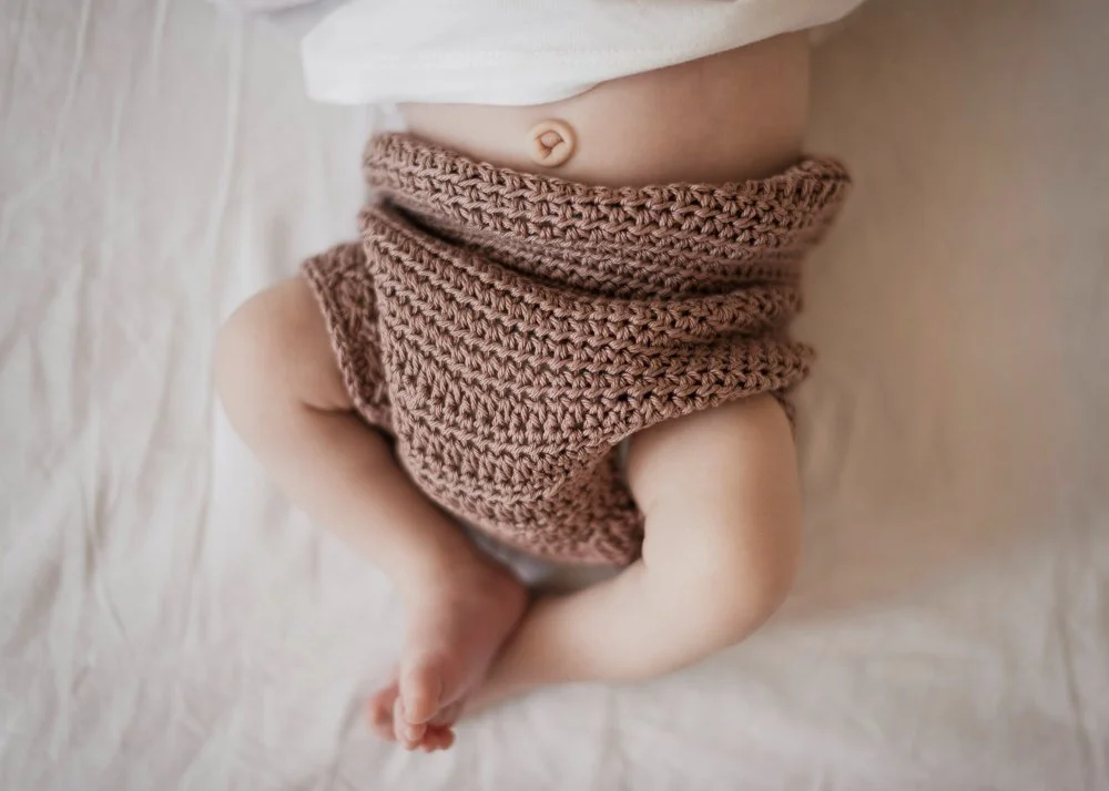 Close-up of a baby's lower abdomen, legs, and feet, wearing a brown knitted diaper cover, lying on a soft, light-colored surface.