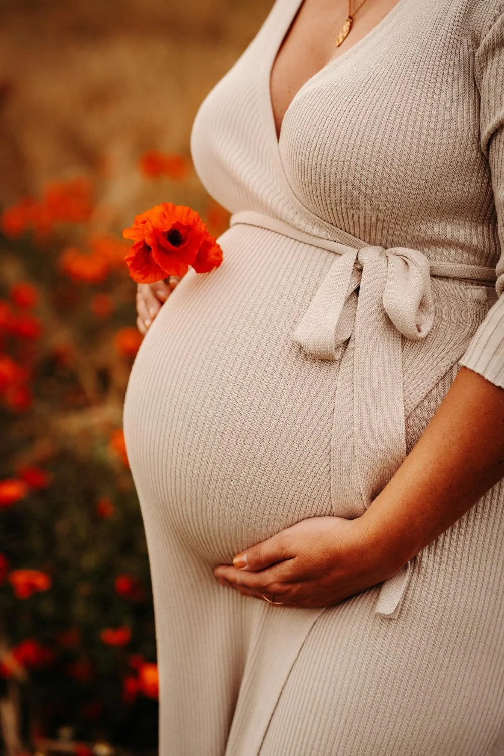 A pregnant woman in a beige ribbed dress holding a red poppy flower, outdoors with blurred red flowers in the background.