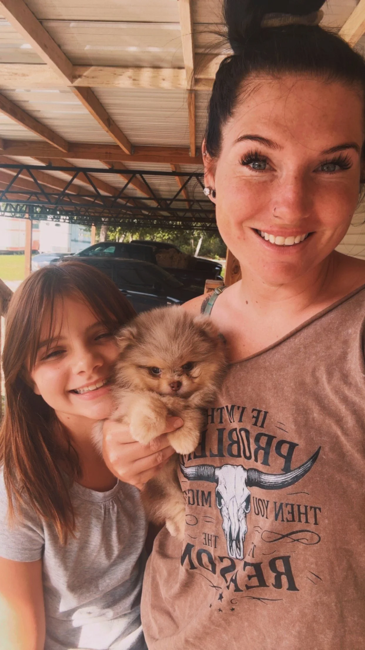 A woman and a girl smiling while holding a fluffy Pomeranian puppy, standing in a partially outdoor, wooden structure area with cars visible in the background.