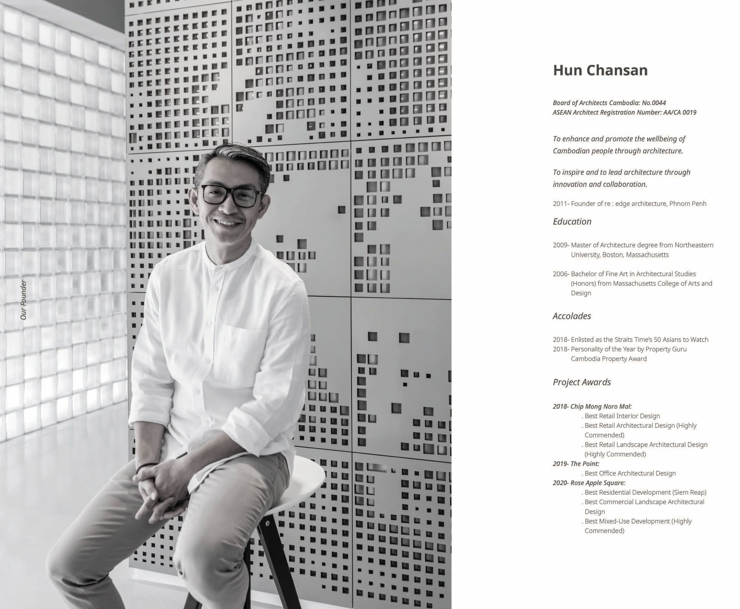A man with glasses and a white shirt sitting on a black chair, smiling, with a modern architectural wall featuring square holes behind him.