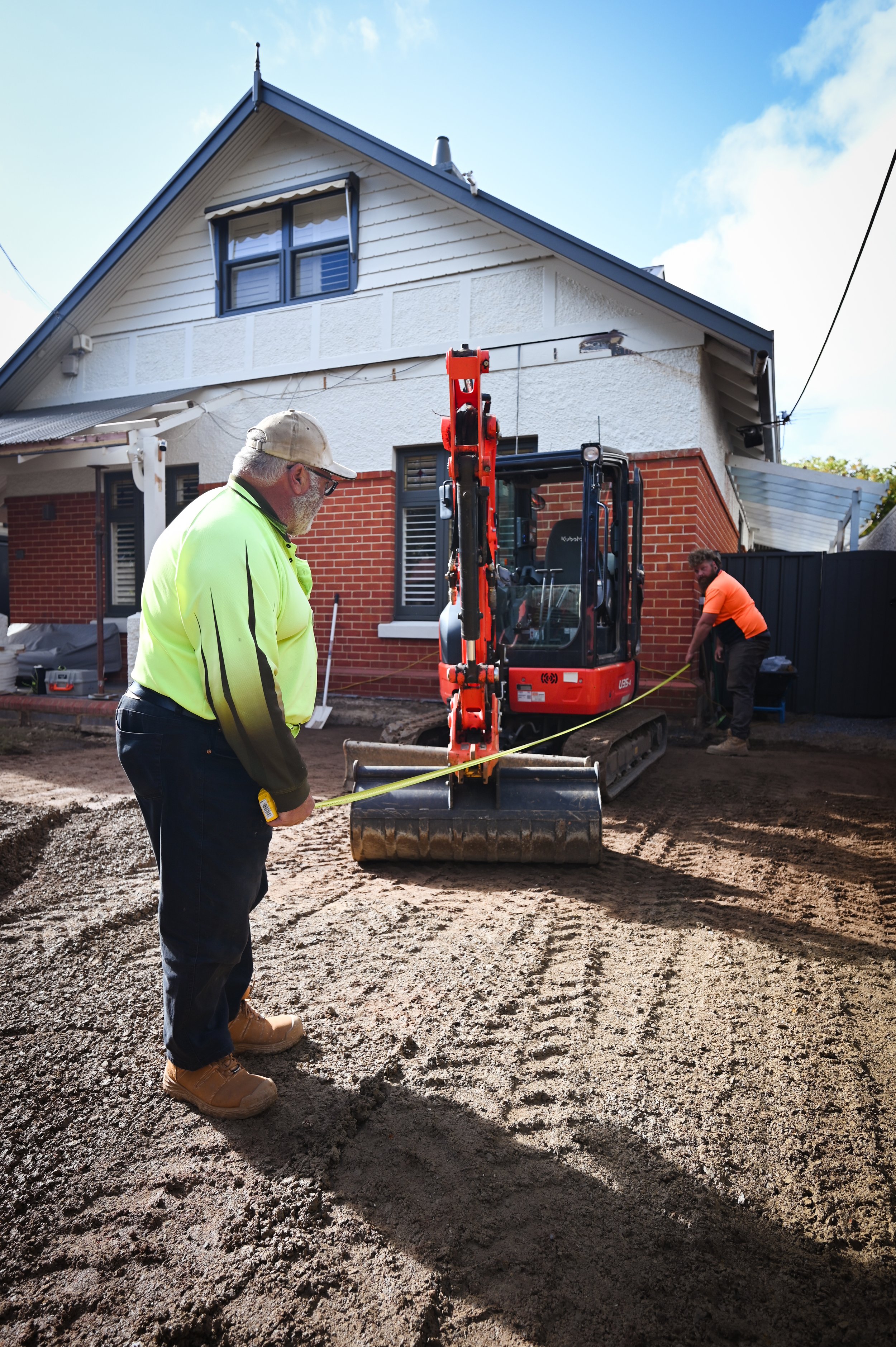 Construction site in front of a residential house with two workers, one in a yellow safety jacket and the other in an orange shirt, operating and supervising a small excavator, with ground excavation work underway.