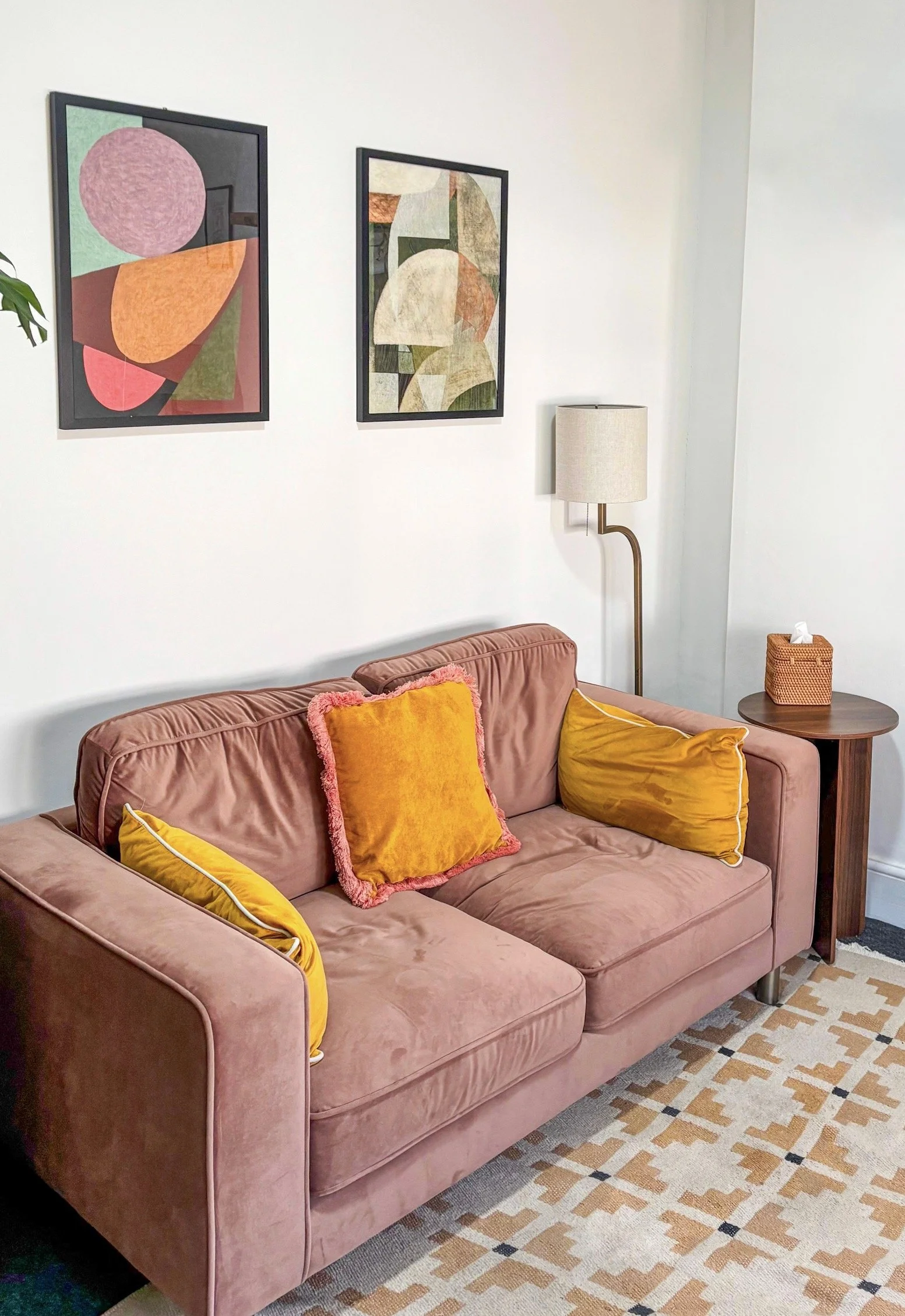 A pink velvet sofa with yellow and pink throw pillows, a wooden side table with a tissue box, and framed abstract art above the sofa, in a cozy living room.