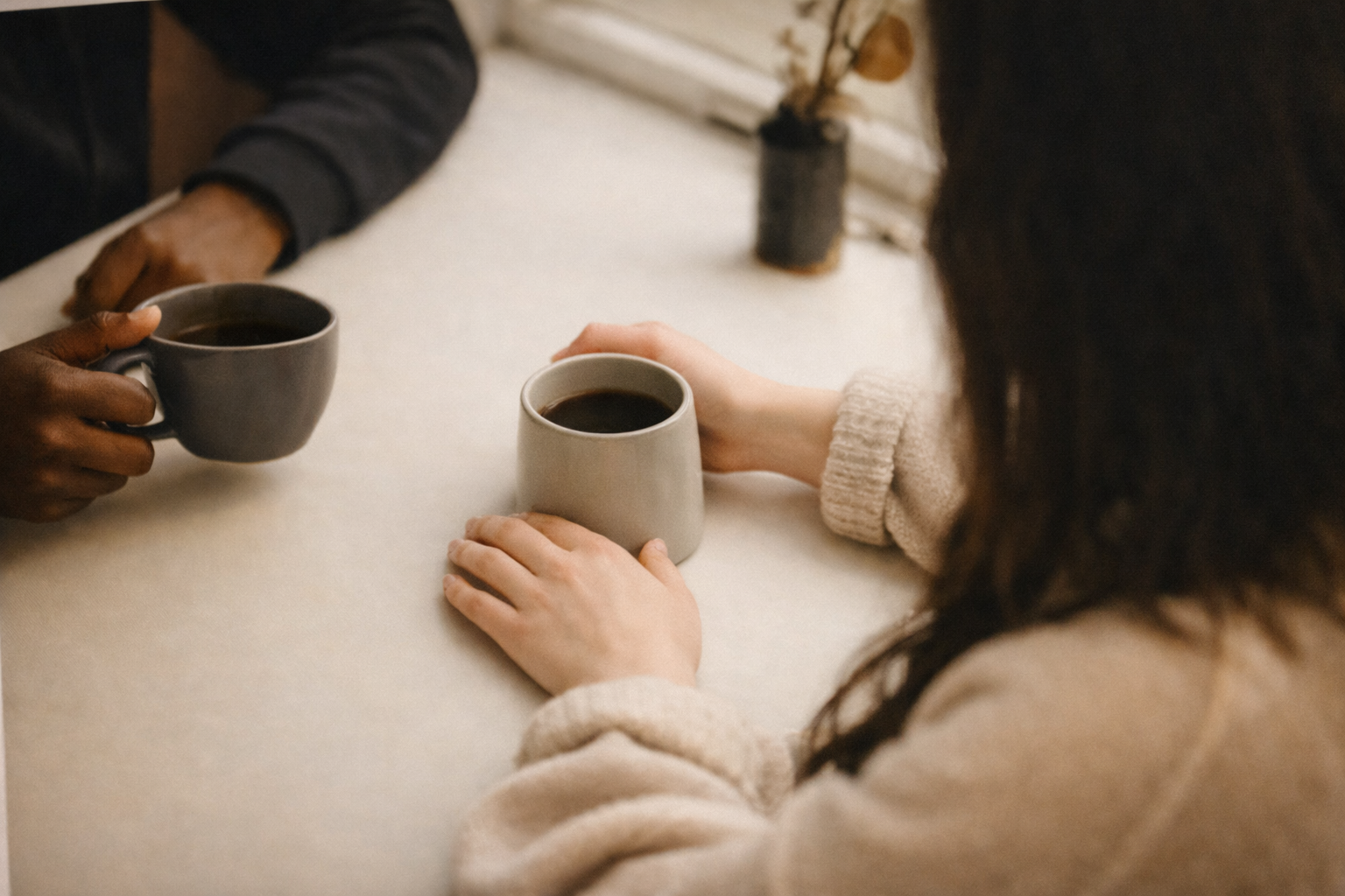 Two women sitting at a table, each holding a coffee mug, with a small, minimalist decor item on the table.