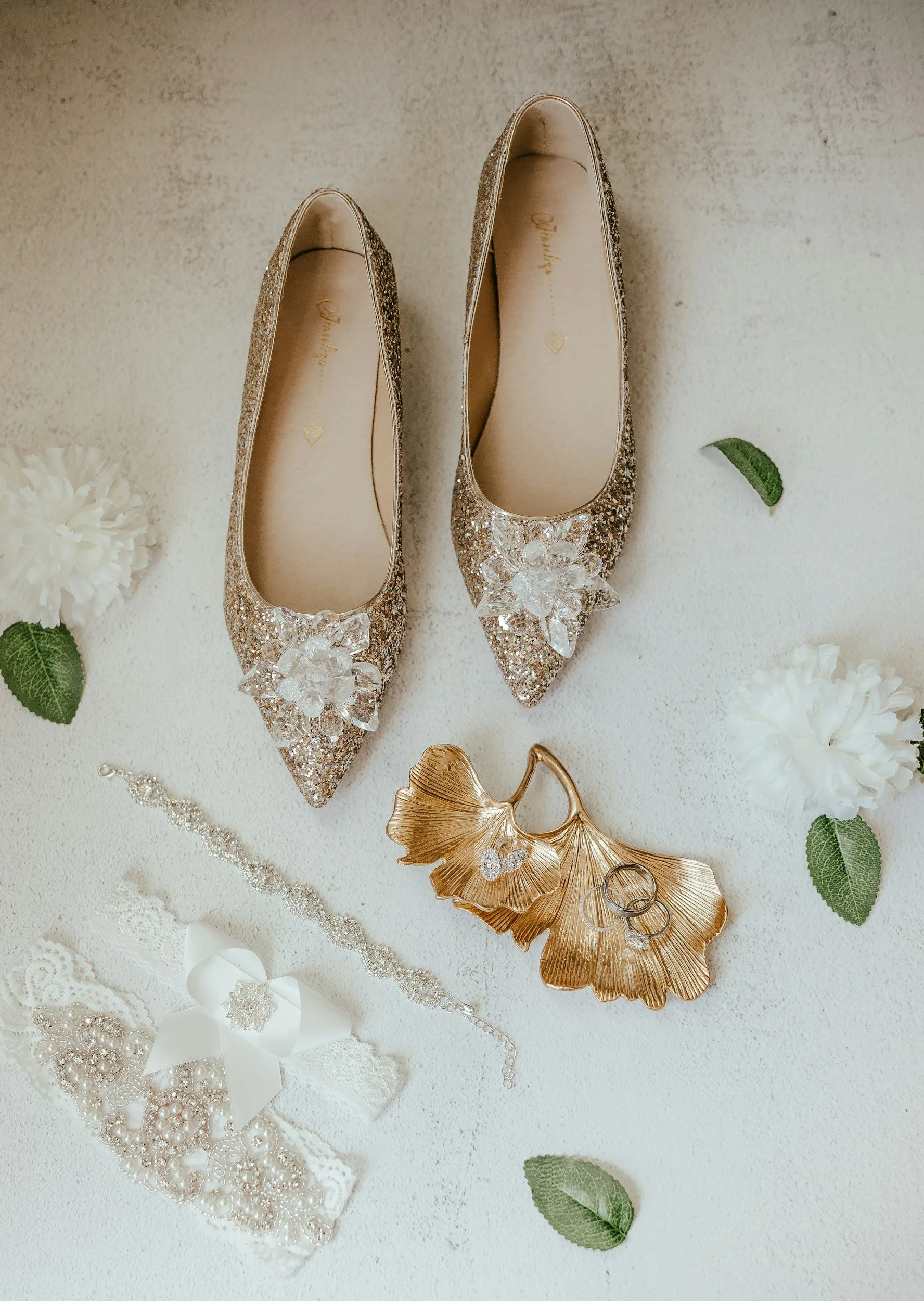 A pair of glittery champagne-colored pointed-toe flats with decorative floral embellishments on the toes, surrounded by white flowers, green leaves, and jewelry on a white textured surface.