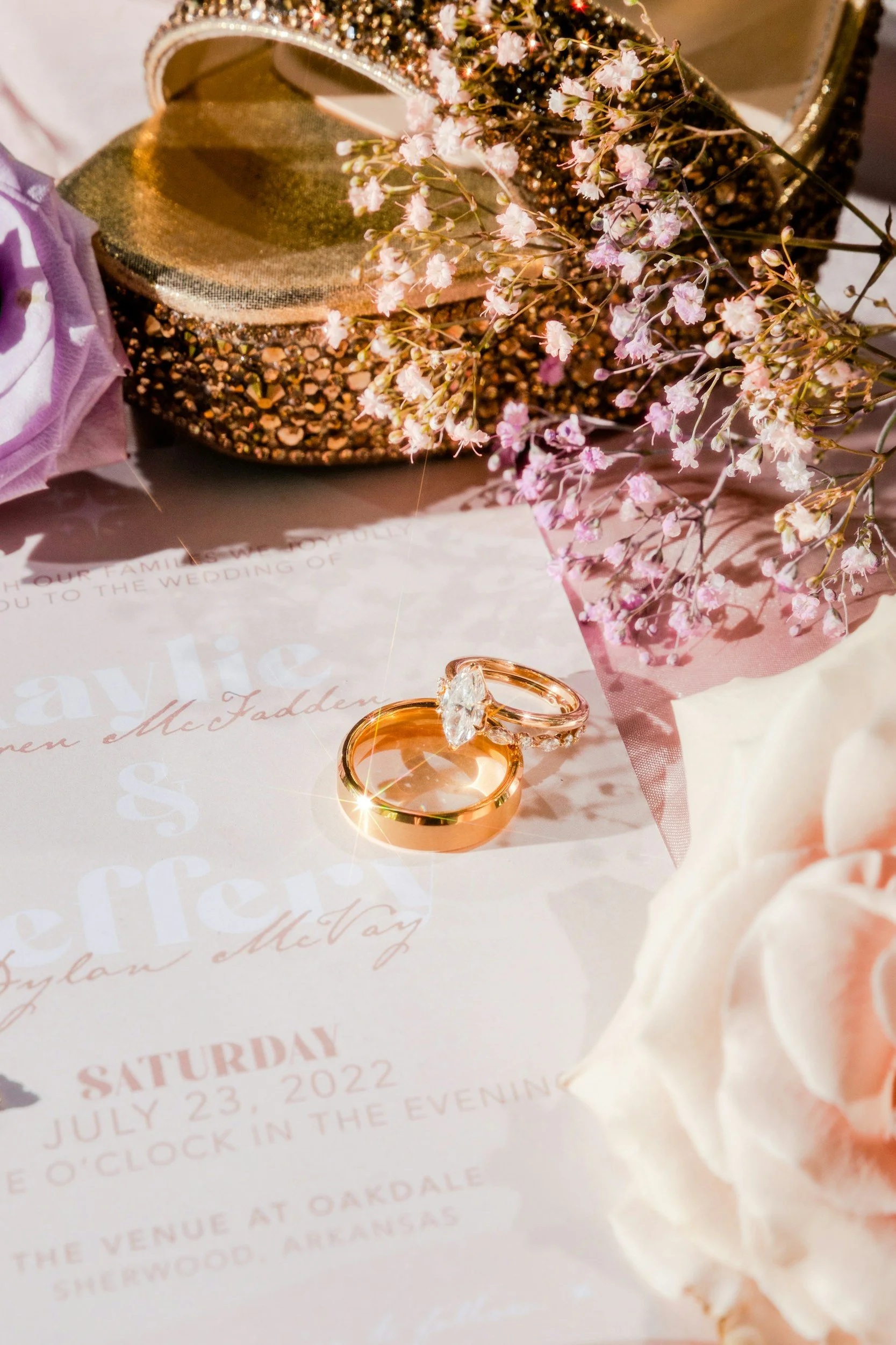 Close-up of wedding rings, pink baby's breath flowers, a glittery gold clutch purse, and a side mirror on a wedding invitation with pink and gold text.