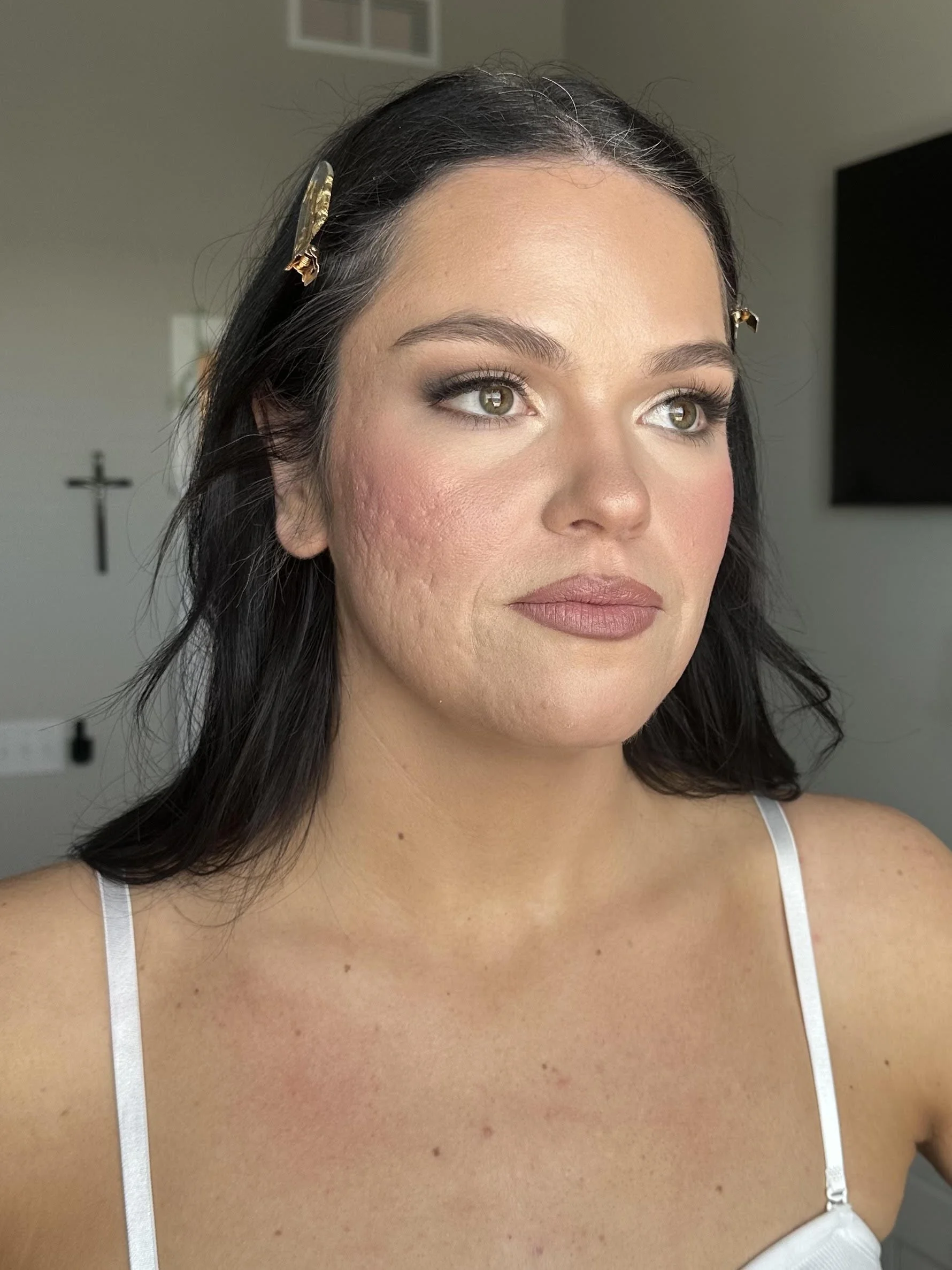 A woman with dark hair, light makeup, and wearing a white top with thin straps is shown in a close-up portrait. Her face has some acne and blemishes, and she has green eyes. natural skin. real skin. unedited makeup. makeup. 