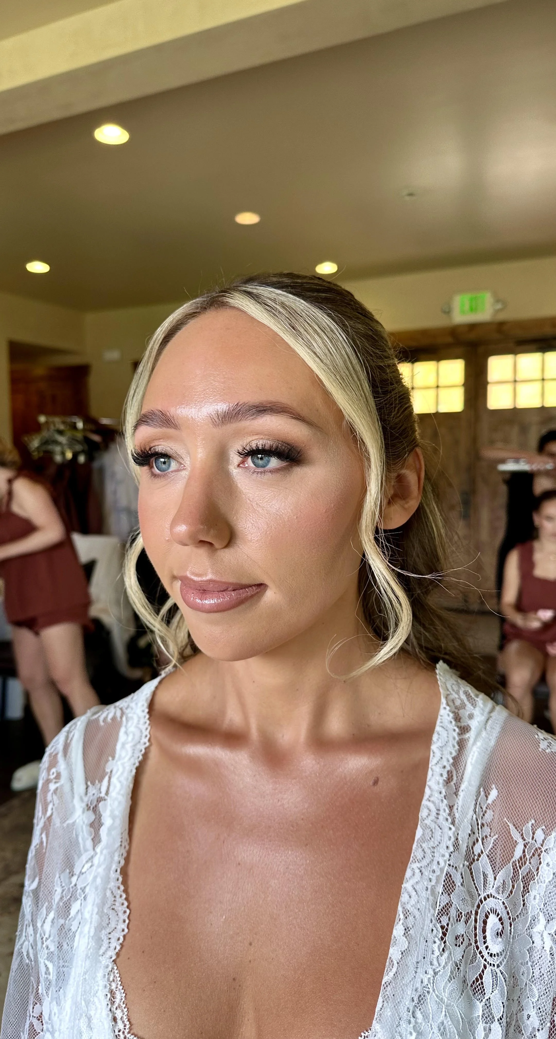A close-up portrait of a woman with blonde hair styled in loose curls, wearing makeup, and a white lace dress, in a room with wooden walls and other women in matching dresses in the background.