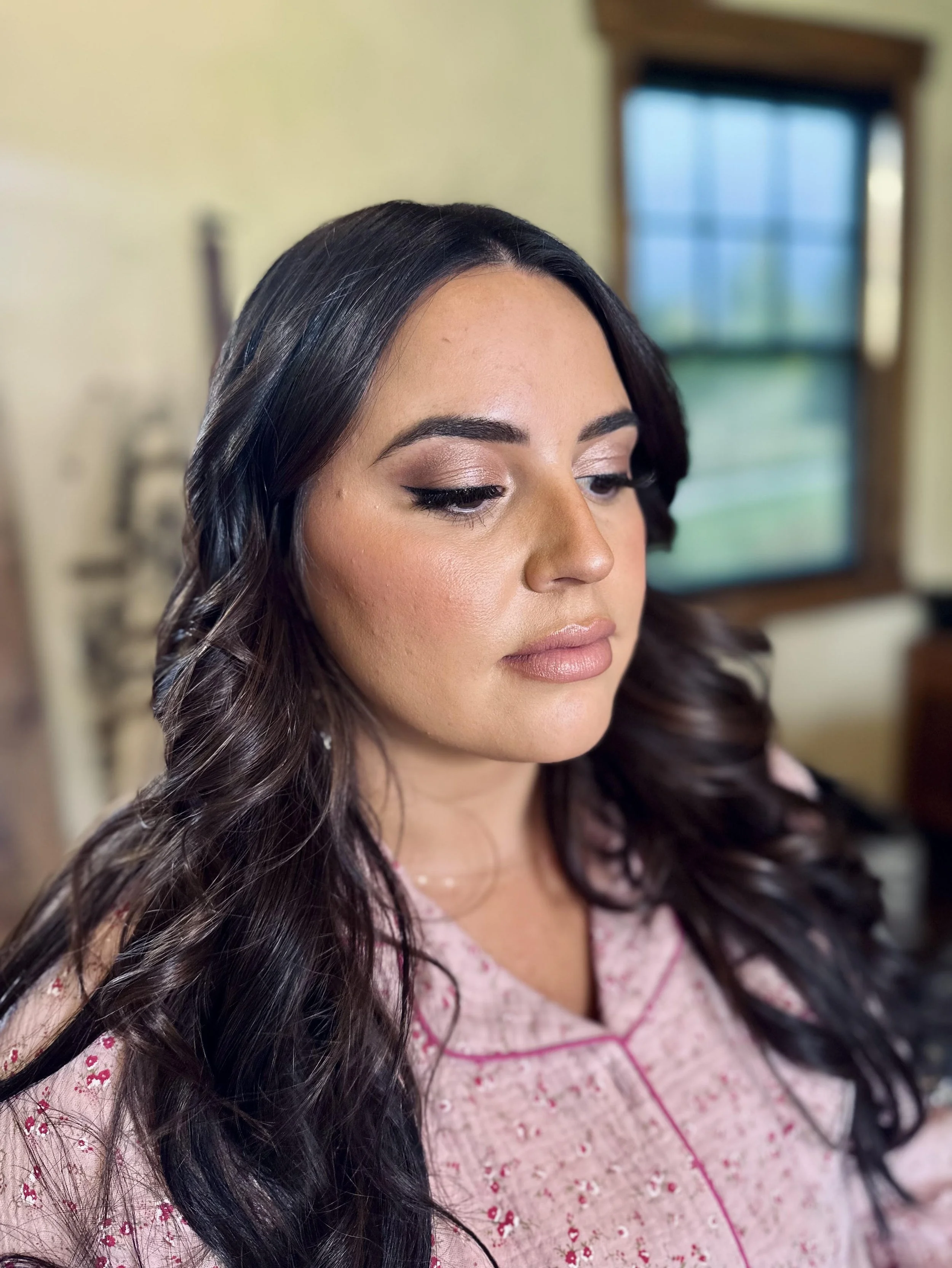 A woman with long, dark, curled hair and makeup, wearing pink and white embroidered pajamas, looking downward, indoors near a window.