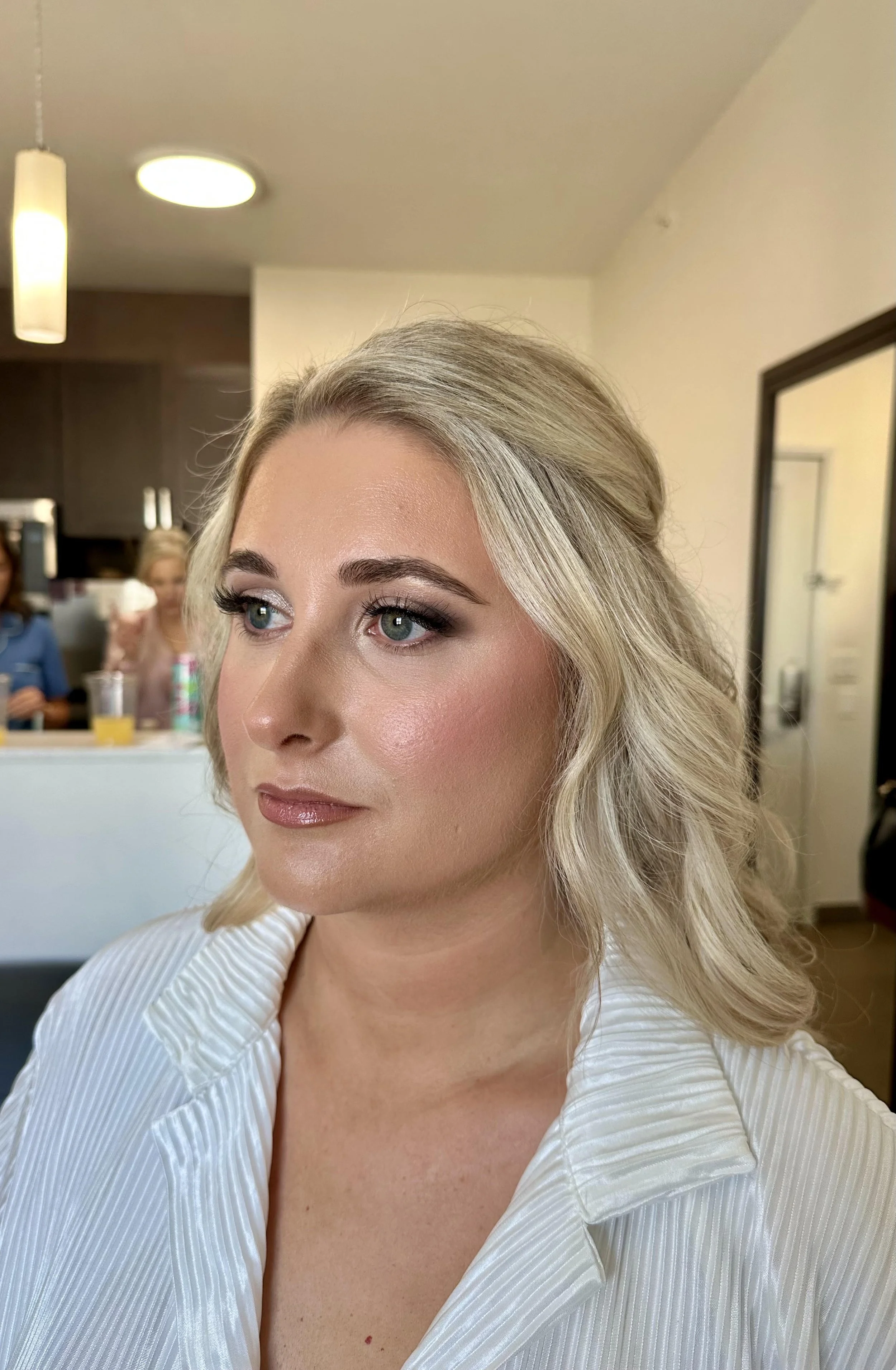 A woman with blonde hair styled in soft waves, light makeup, and wearing a white, pinstripe blouse with a V-neck, is looking to the side in an indoor setting that appears to be a dressing room or backstage area.