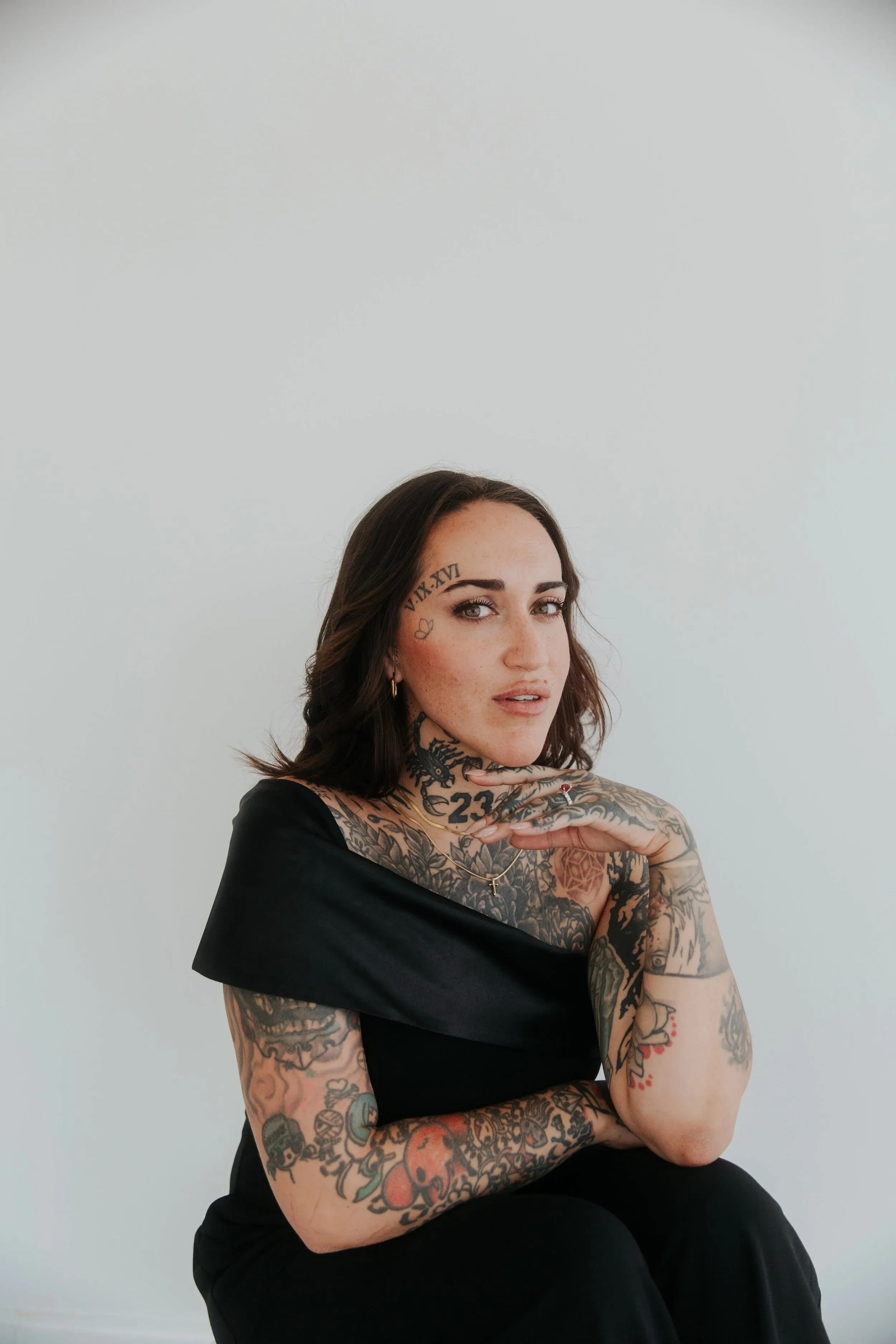 A woman with dark brown hair and tattoos on her face, arms, and chest, wearing a black off-shoulder top, sitting against a plain light gray background.