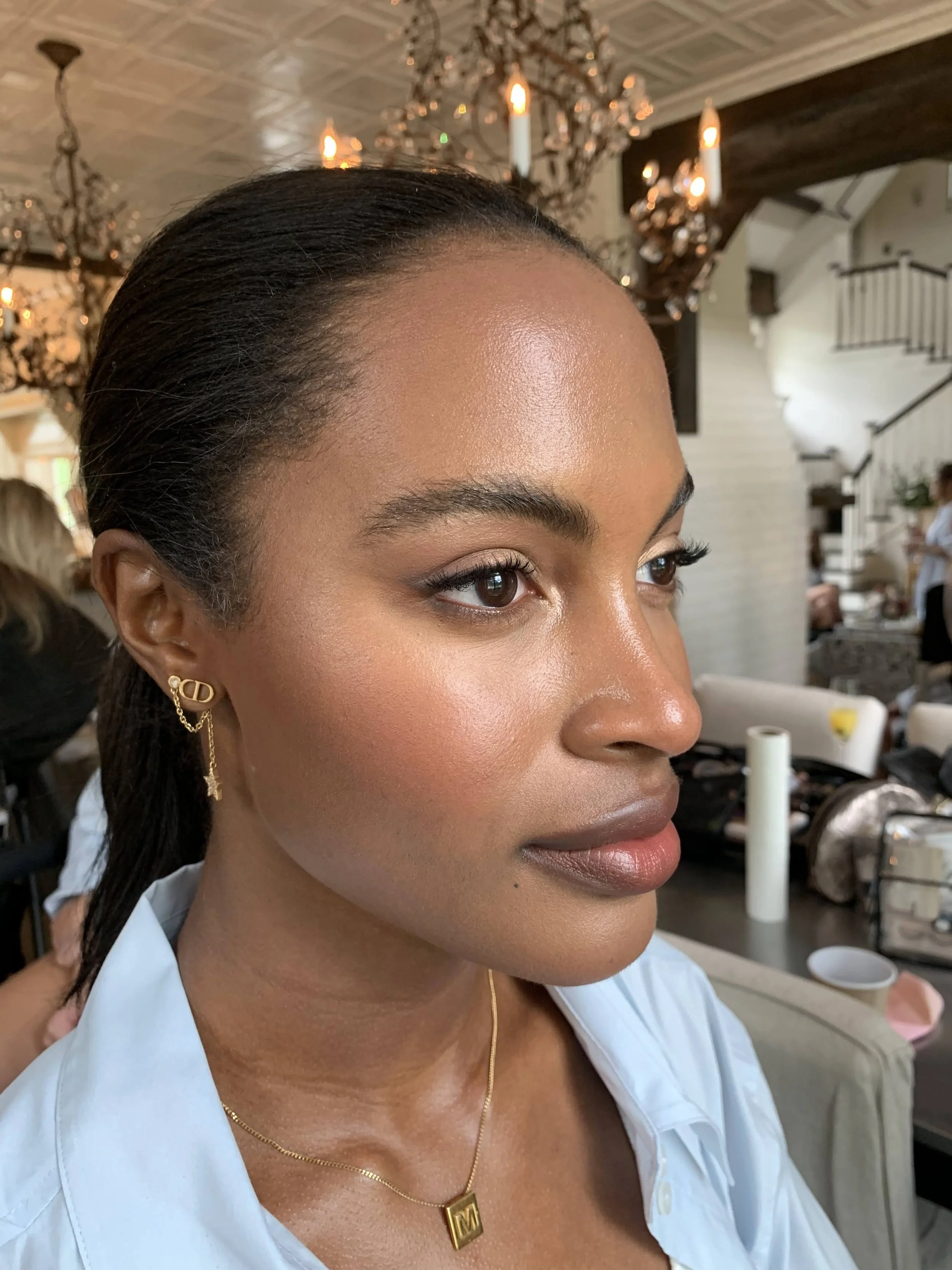 Close-up of a woman with dark hair pulled back, wearing gold jewelry, in a well-lit interior space with chandeliers and a staircase in the background. Beautiful makeup on a black woman. natural makeup. makeup for deeper skin tones.