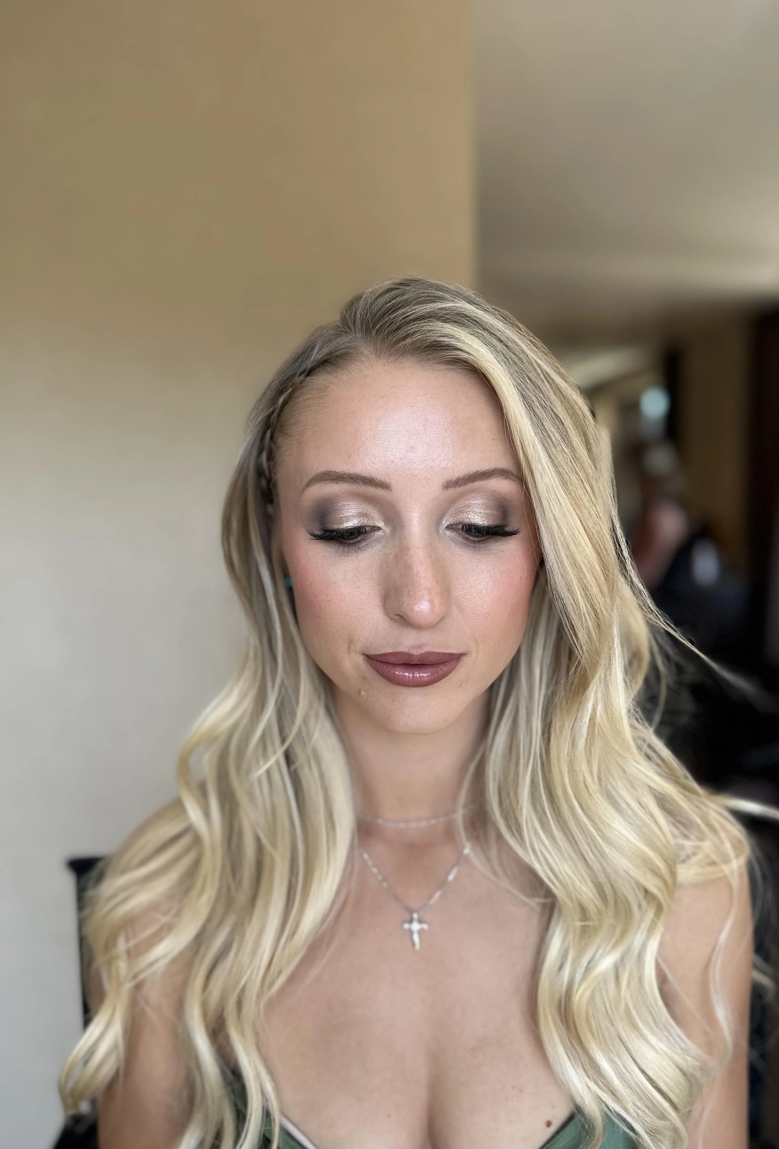 A young woman with long blonde hair styled with loose waves and a small braid on the side, is wearing eye makeup and a silver cross necklace, looking downward in an indoor setting.