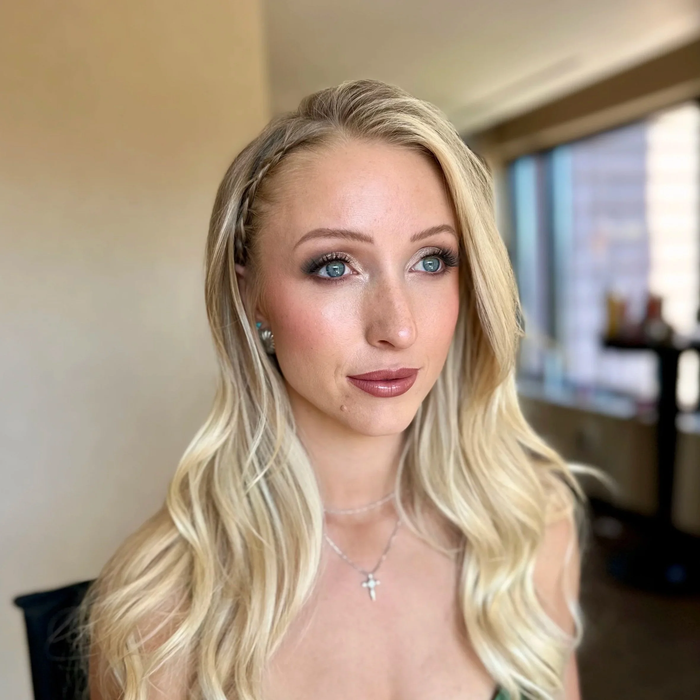 Close-up of a woman's face with long blonde hair, blue eyes, and wearing a necklace with a cross pendant, standing indoors near a window. Beautiful makeup. real skin. natural skin. glam. bridal glam.