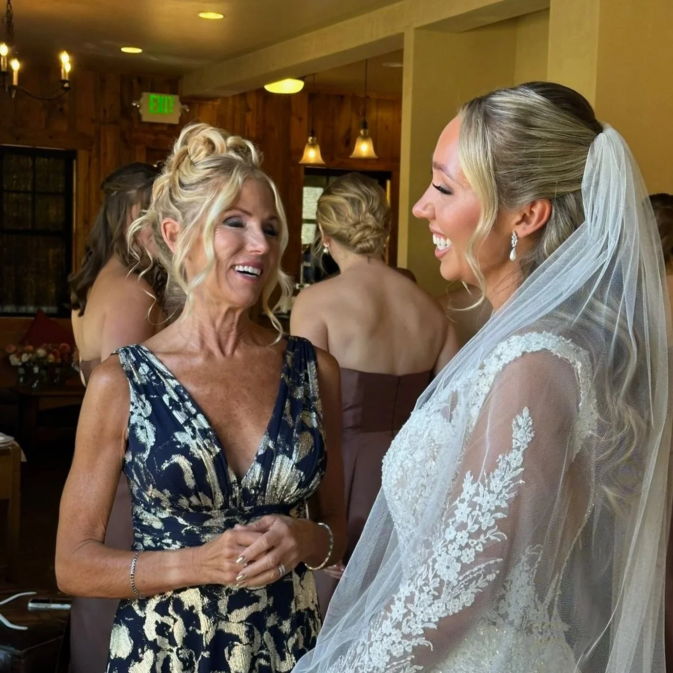 A bride with a veil and lace dress smiling at an older woman in a blue and gold dress, both sharing a joyful moment at a wedding reception. bridal glam. Bride. Glam makeup. makeup artist.