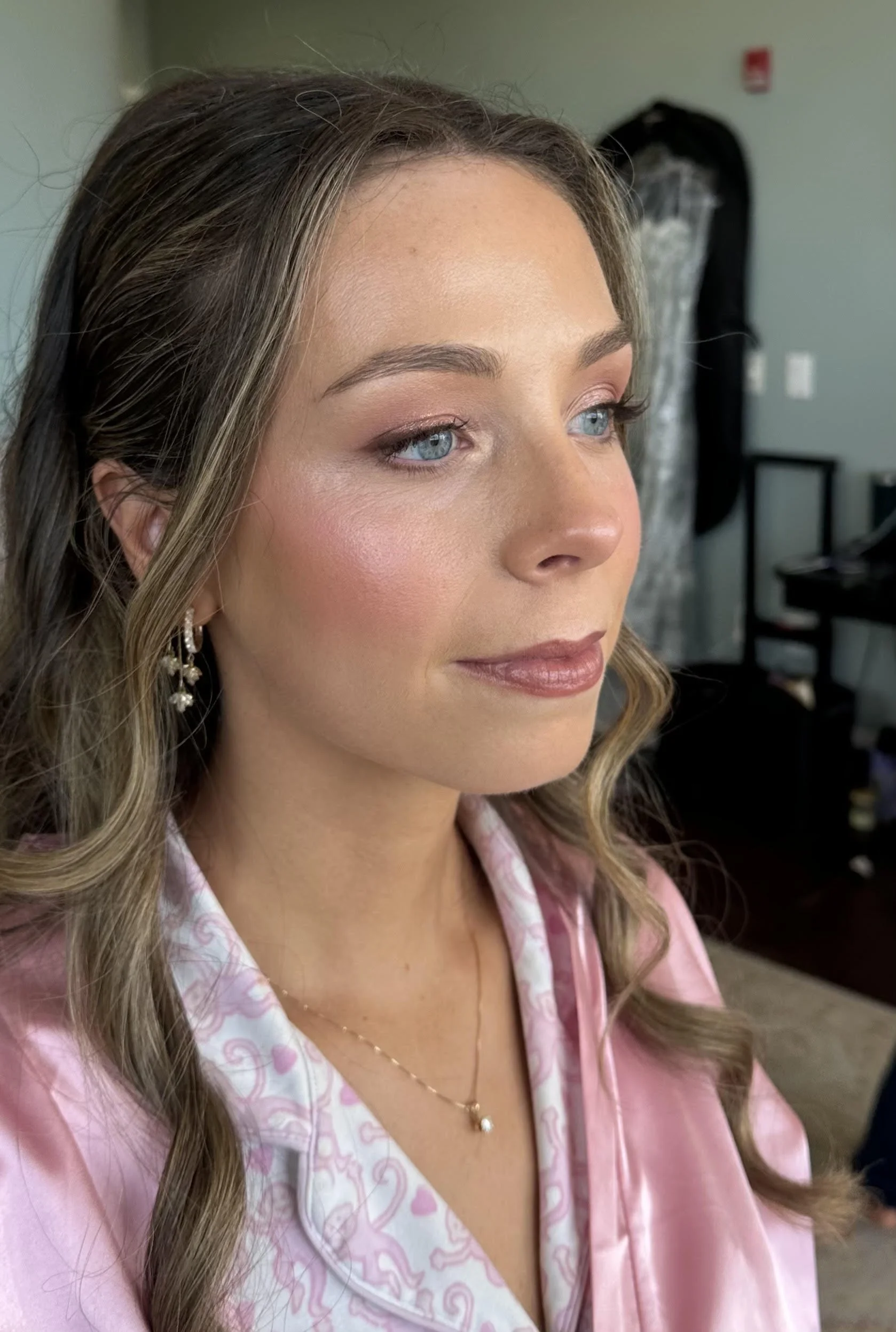 A woman with wavy, light brown hair and blue eyes, wearing pink pajamas with heart and swirl patterns, earrings, and a necklace, standing in a room with a green wall and a black luggage bag in the background.