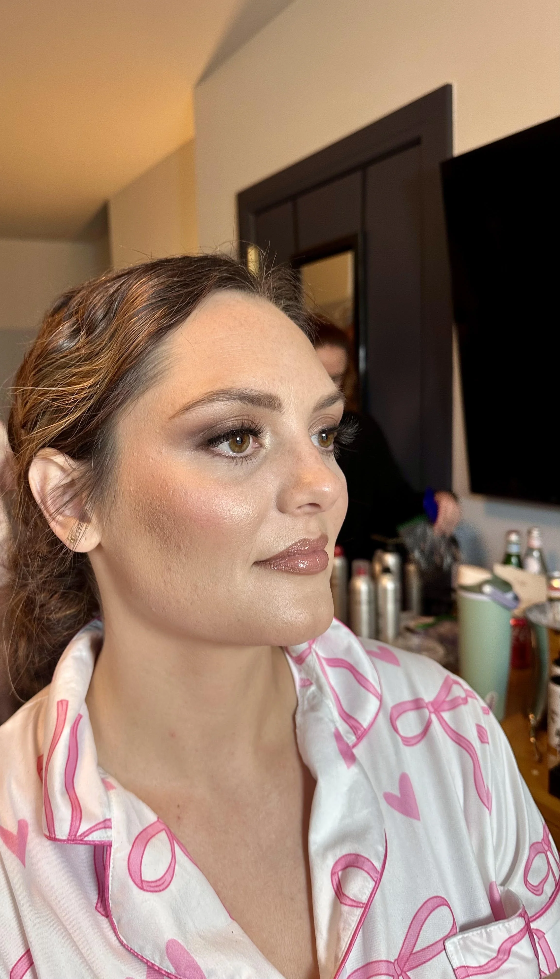 A woman with brown hair and makeup, wearing pink and white pajamas with heart patterns, sitting in a room with a mirror and beverages on a table. Bridesmaid makeup. Bridesmaid. Hair and makeup. Bridal makeup. Bride. Makeup 