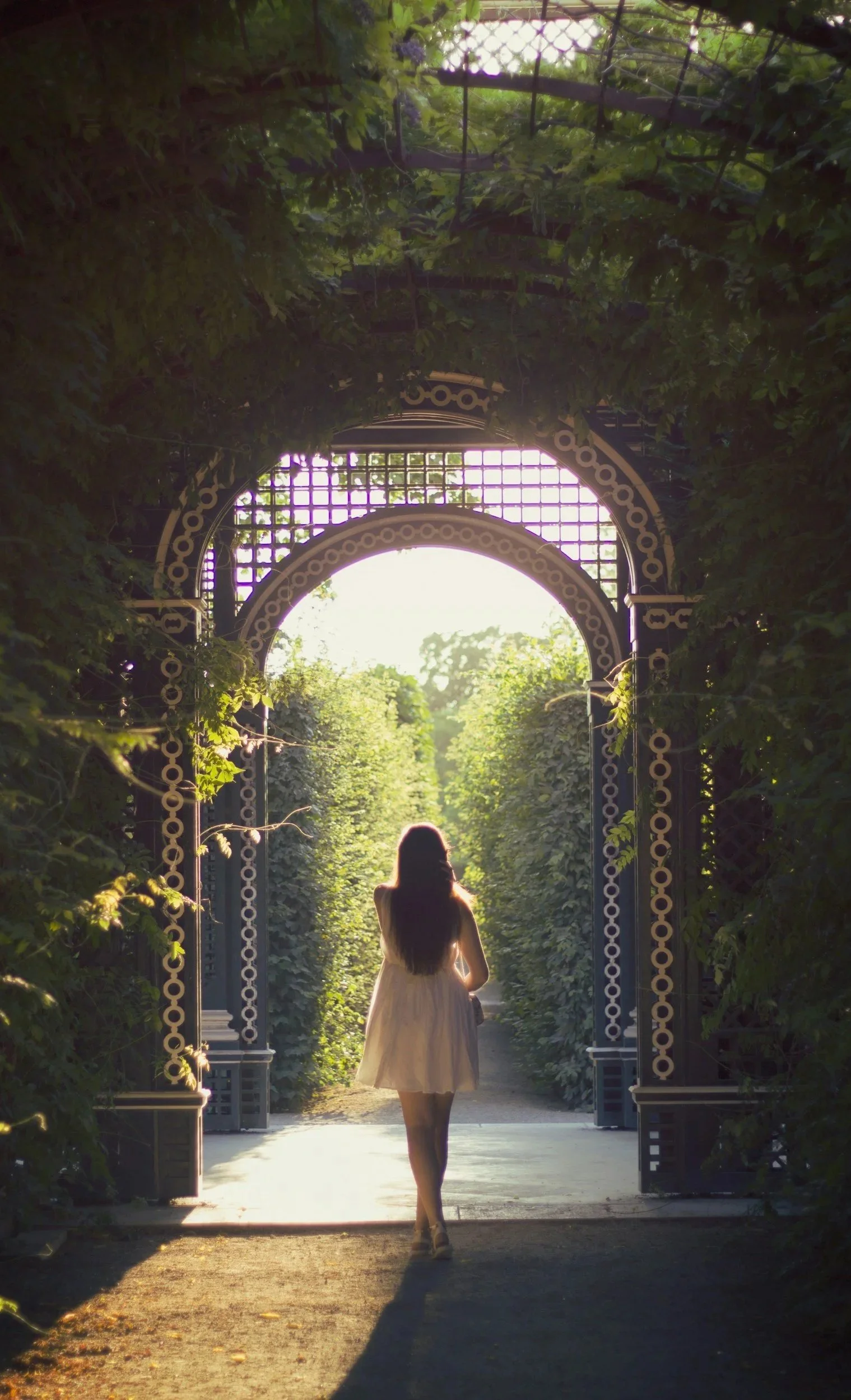 A woman with long hair wearing a light-colored dress walking through a decorative garden archway with greenery and trees in the background, illuminated by warm sunlight.