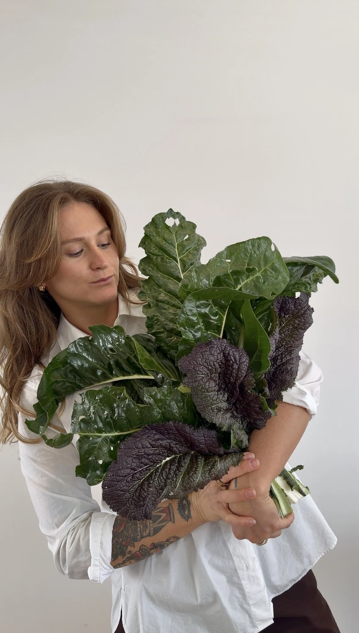 A woman with light brown hair wearing a white shirt is holding a large bouquet of leafy greens, including kale and other dark green and purple leaves, against a plain white background.
