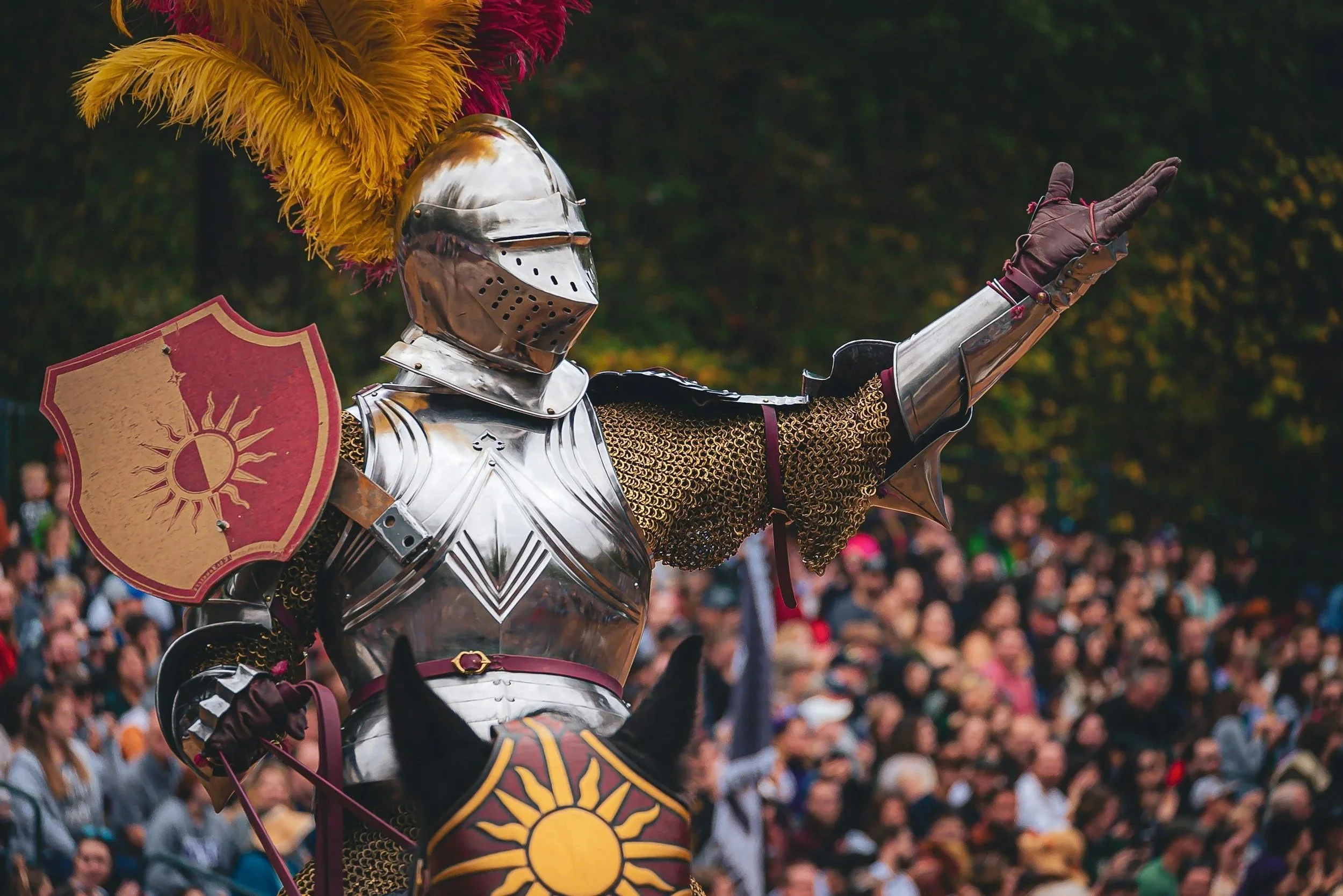 A person dressed as a medieval knight in shiny armor, holding a red shield with a sun emblem, and wearing a gold and red feathered hat, at an outdoor event with a crowd of spectators in the background.