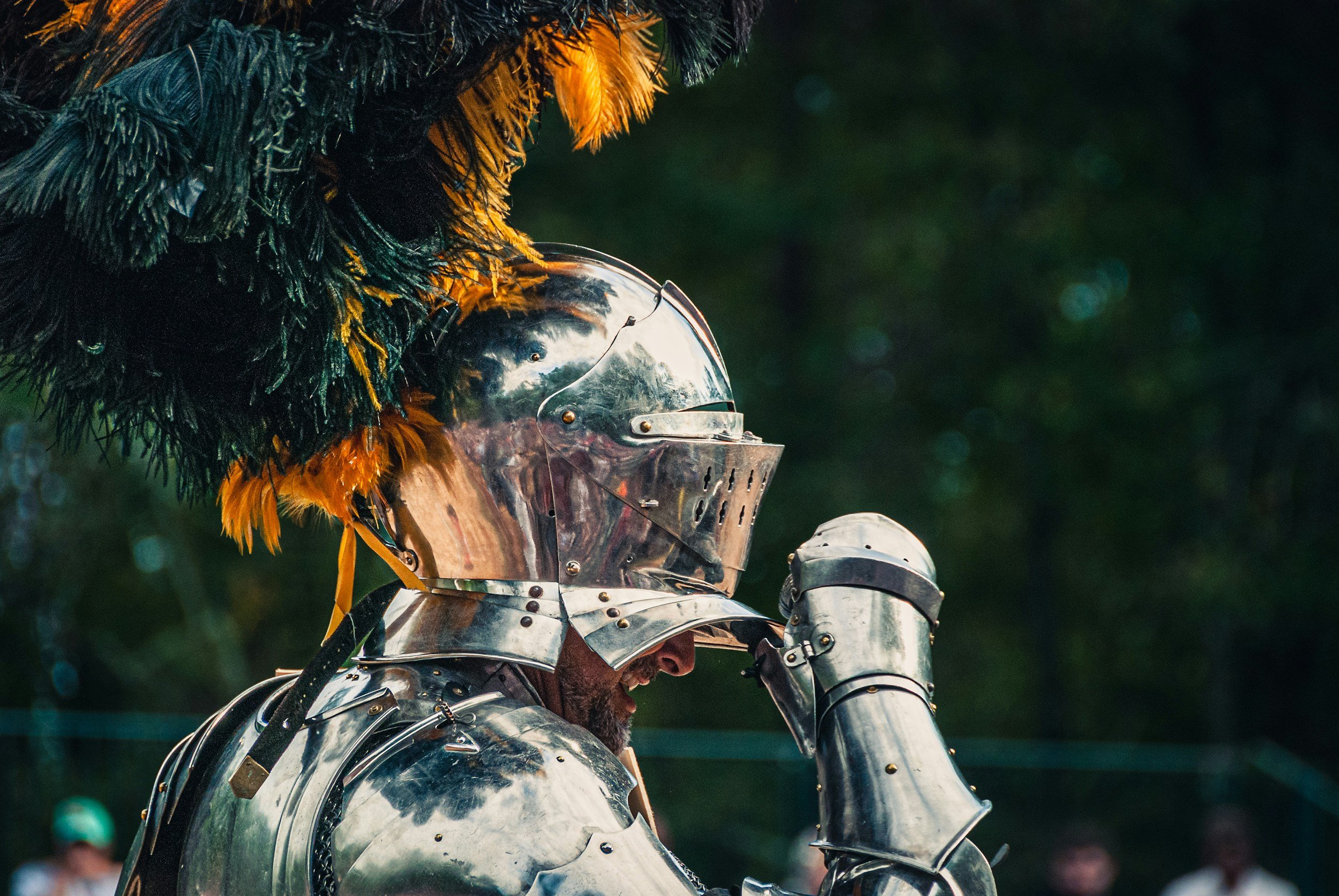 A person dressed as a medieval knight in shiny silver armor is shown adjusting a helmet with a visor. The knight is wearing a feathered hat with green, orange, and yellow feathers. The background is blurred with bokeh effects, suggesting an outdoor setting.