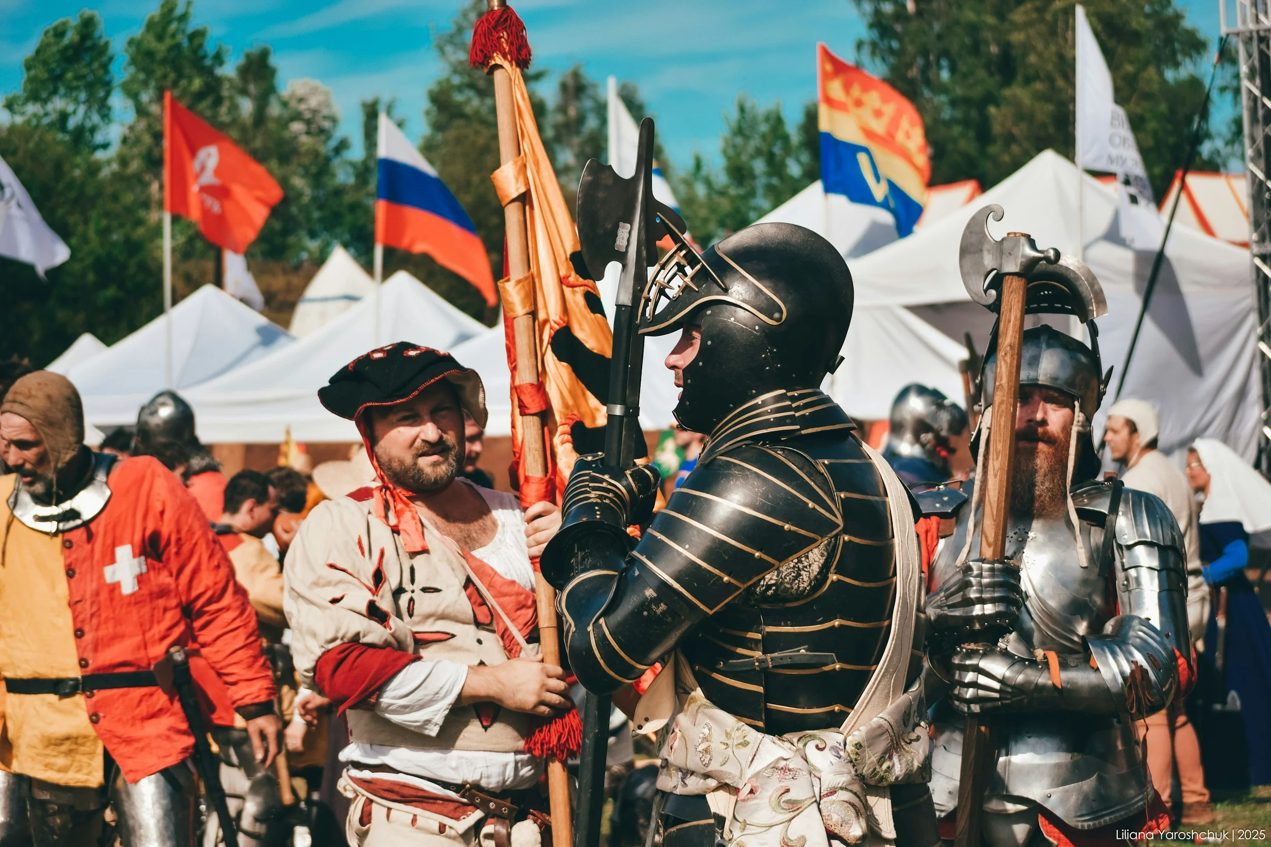 Scene of historical reenactors dressed in medieval armor and period costumes at a festival, with flags and tents in the background.