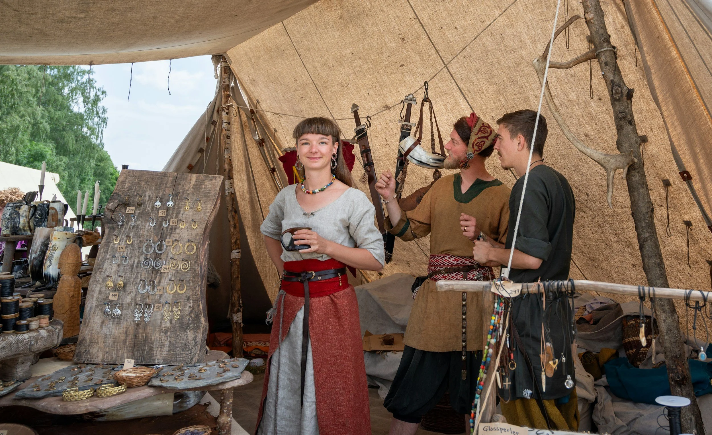 Three people are inside a tent at a market stall displaying handmade jewelry and accessories. The woman in the center has long brown hair and is wearing a gray top with colorful jewelry, smiling and holding a mug. The men on the right are dressed in medieval or Viking-style clothing, one with a red head covering, and are engaged in conversation with the woman. The stall has various jewelry items, including earrings and bracelets, on display.