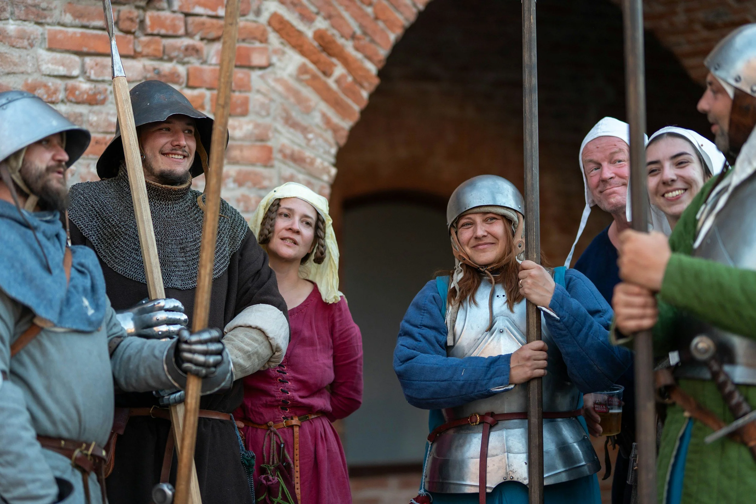 Group of people dressed in medieval costumes, including chainmail, helmets, and armor, standing together outside near a brick building, smiling and holding weapons, possibly preparing for a reenactment or historical event.