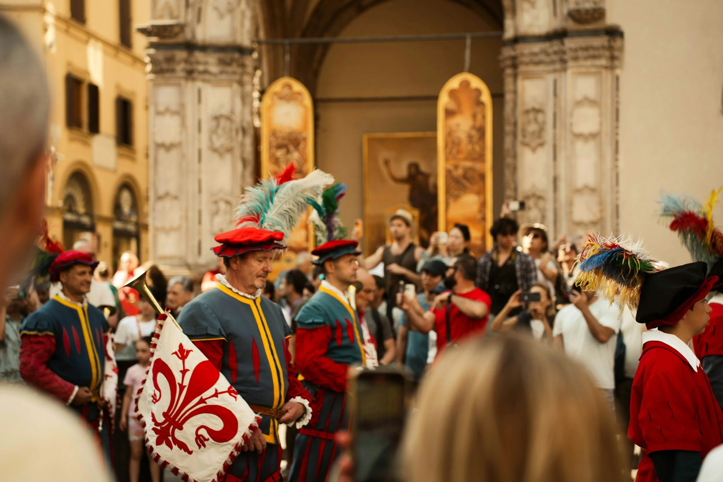 People dressed in medieval costumes with colorful feathered hats and robes, participating in a parade or festival inside a historic building with ornate architecture and religious artwork.