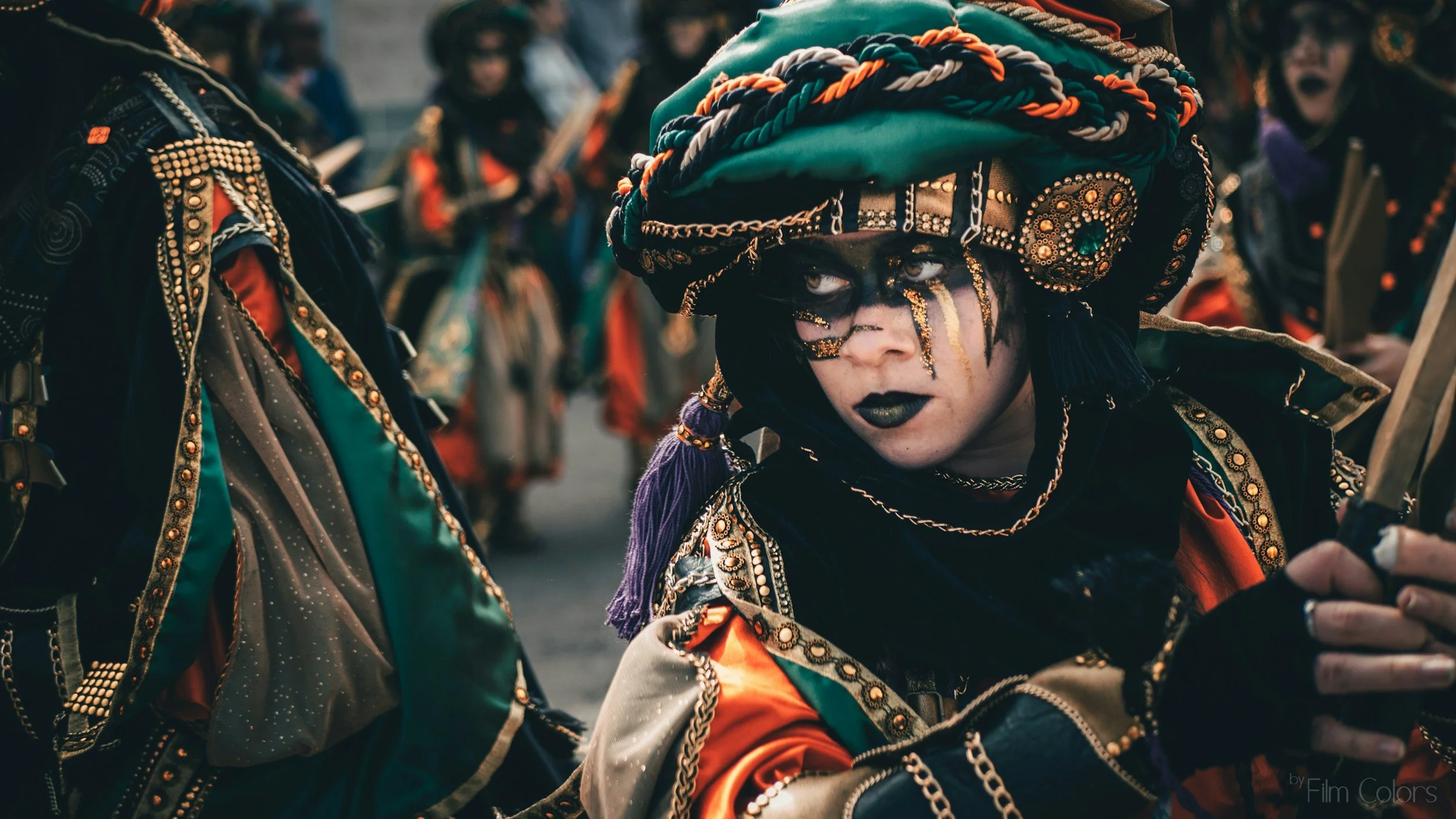 Person with black face makeup and gold accents, wearing a large green, orange, white, and black braided headwrap, dressed in colorful ornate clothing, surrounded by other people in similar costumes, during a festival or parade.