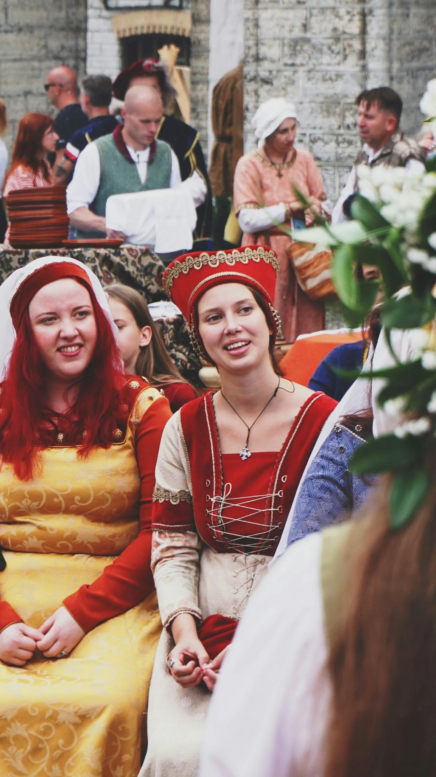 People dressed in traditional costumes attending a cultural event or celebration, with some women wearing red and gold dresses, and others in historical attire, in front of a stone wall.